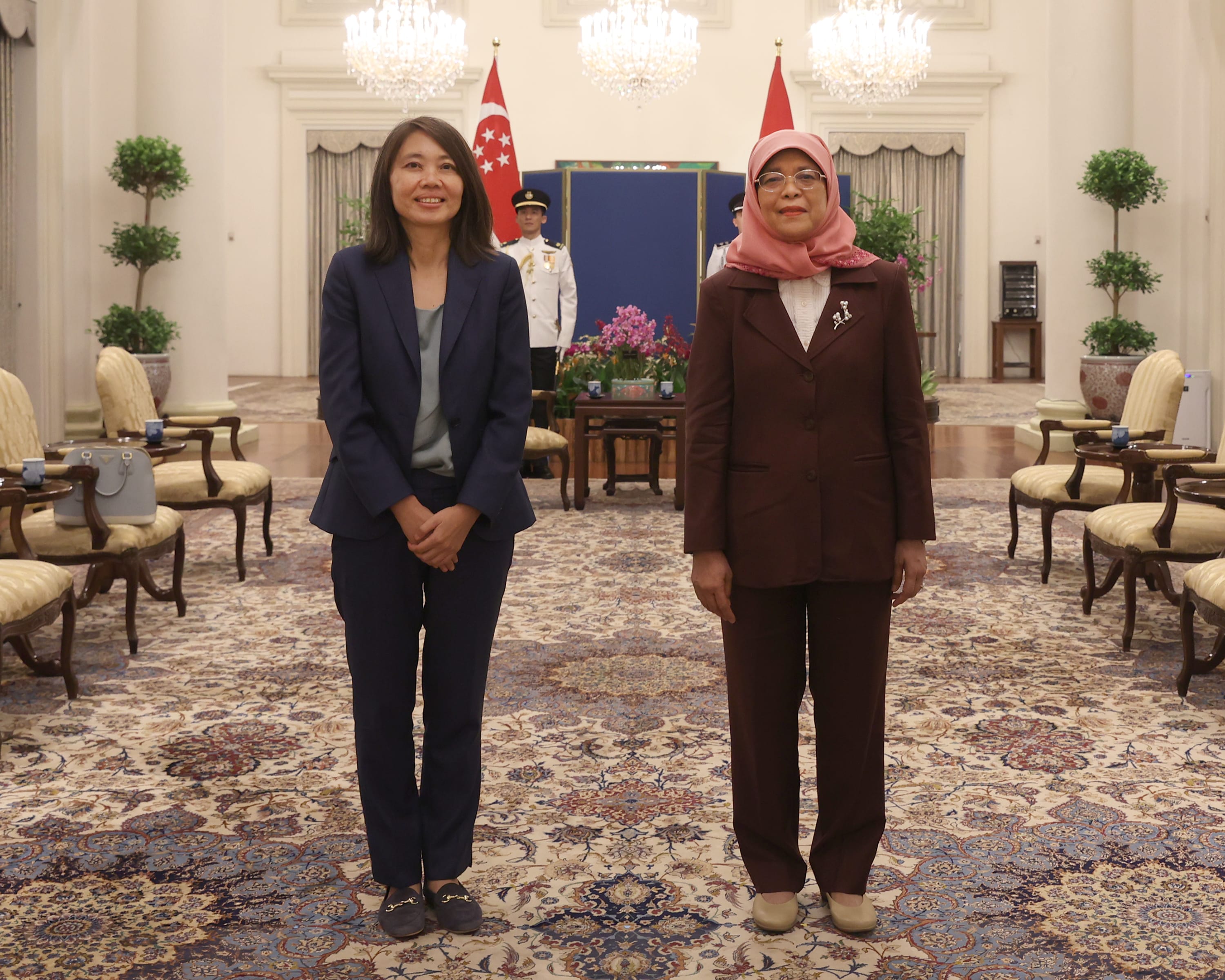 Two women in suits stand in a room with the Singapore flag. One woman wears a hijab.