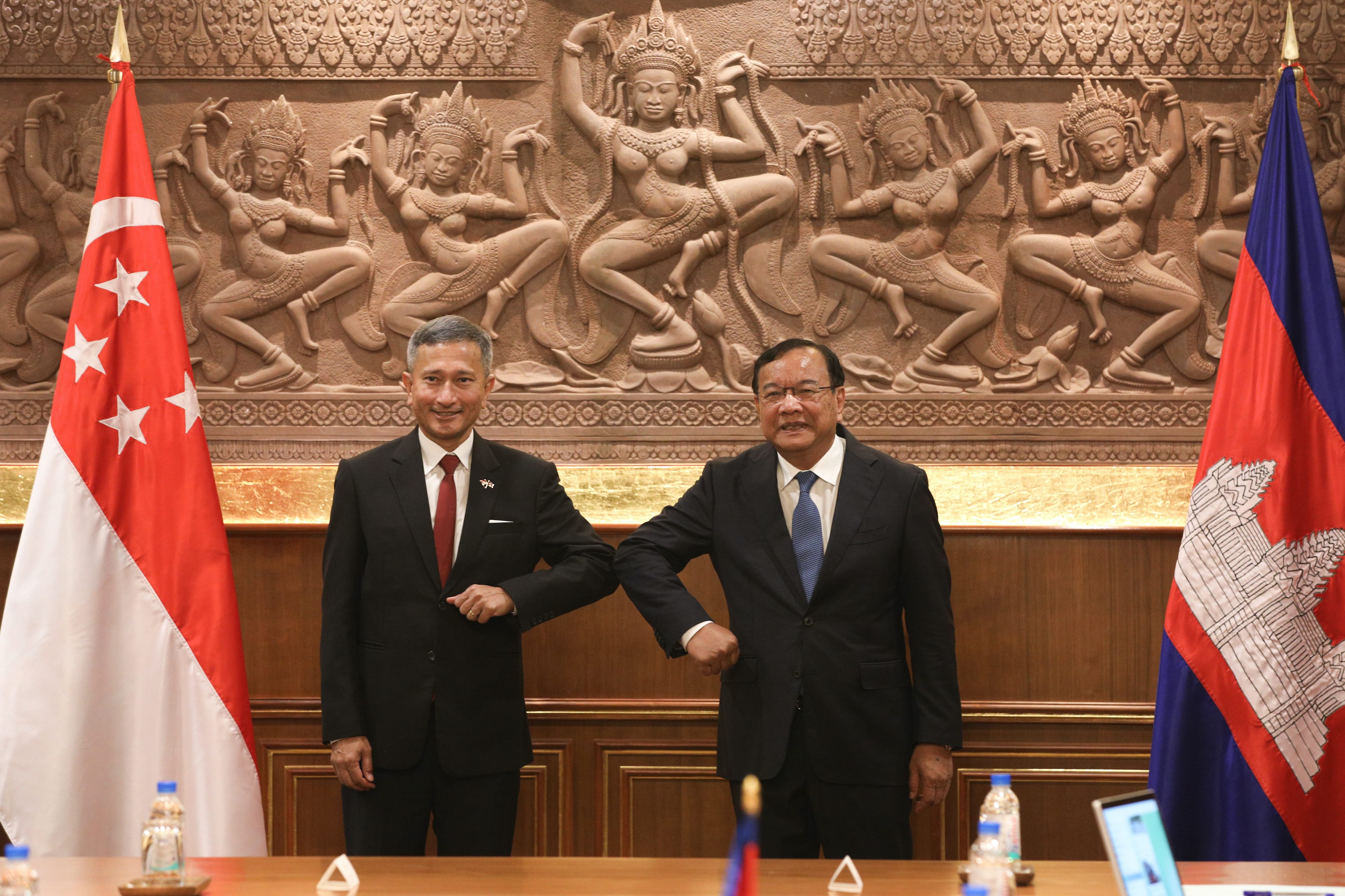 Two men in suits bump elbows between Singapore and Cambodia flags. Carved relief in the background.