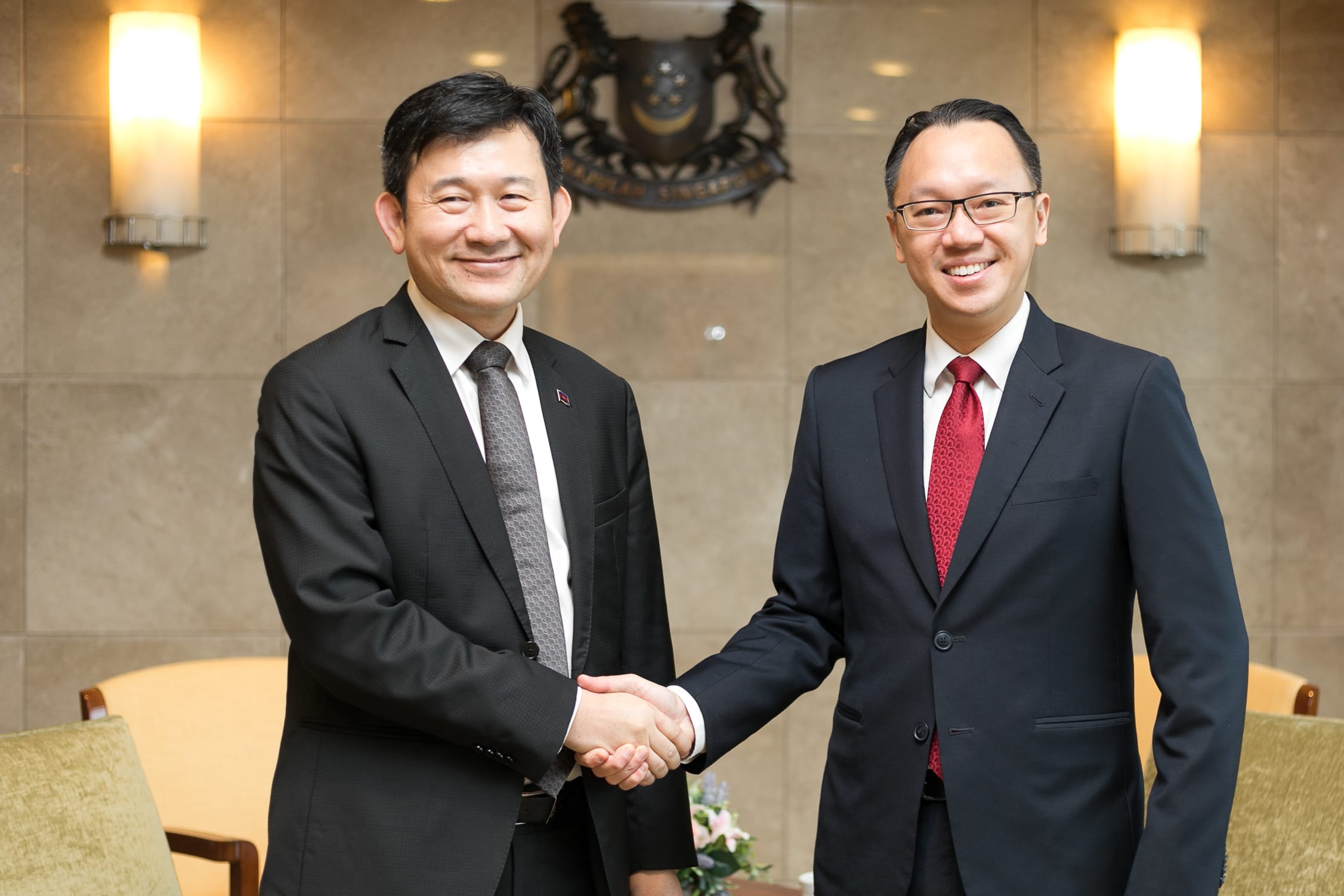 Two men in suits shaking hands in front of a Singapore coat of arms.