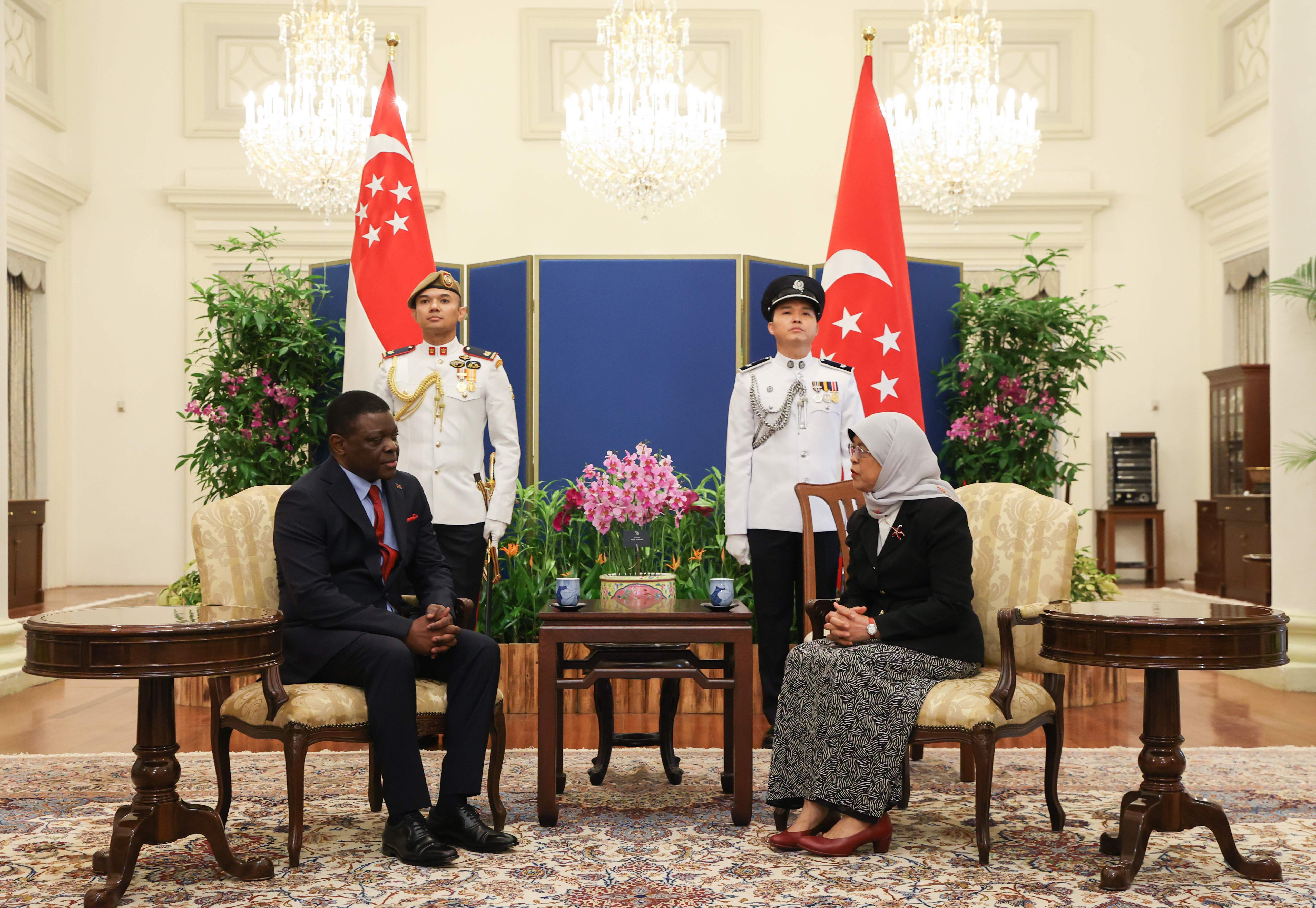 Two people seated between Singapore flags, flanked by guards in ceremonial dress.