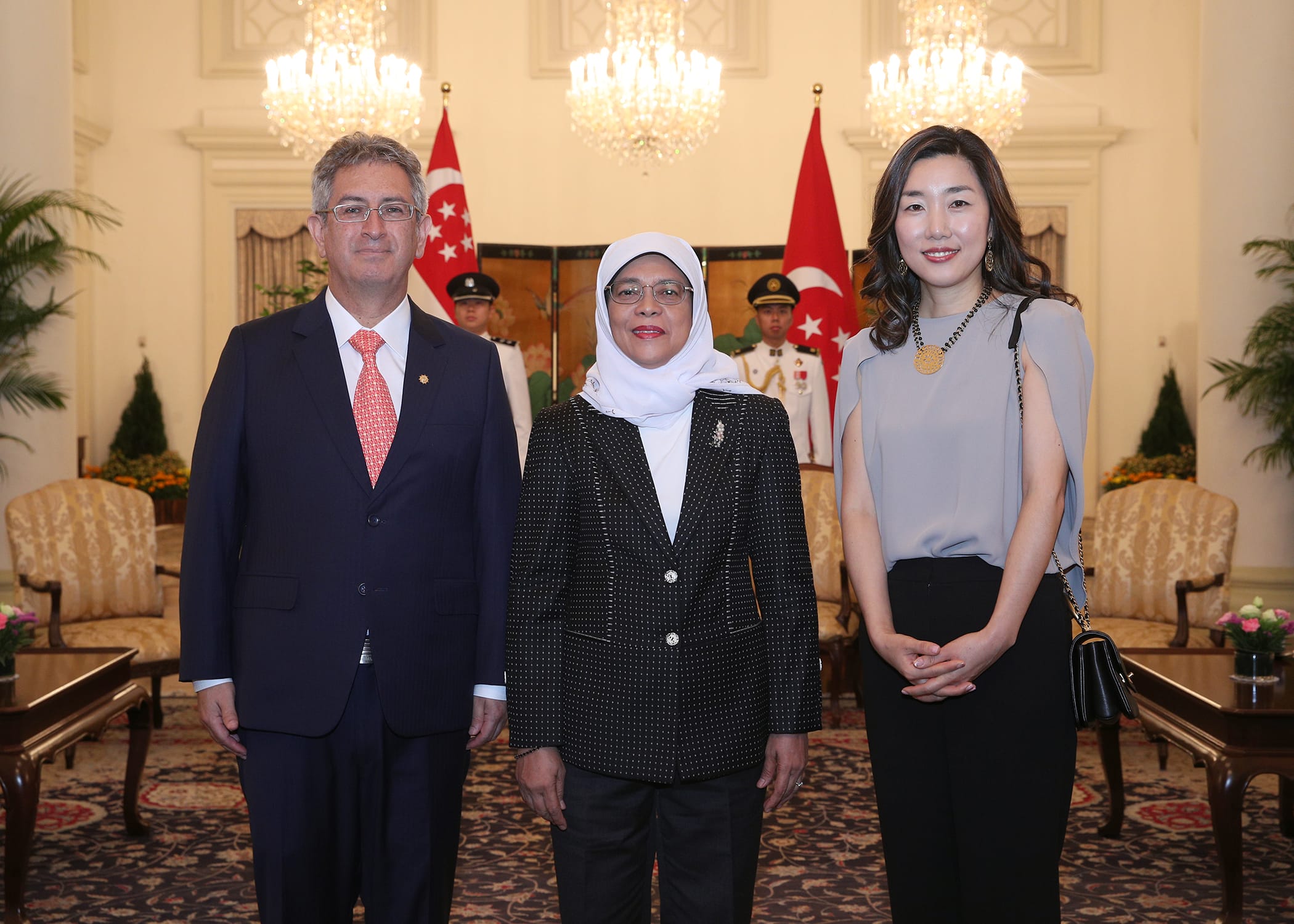 Three people stand indoors before Singapore flags and honor guard.