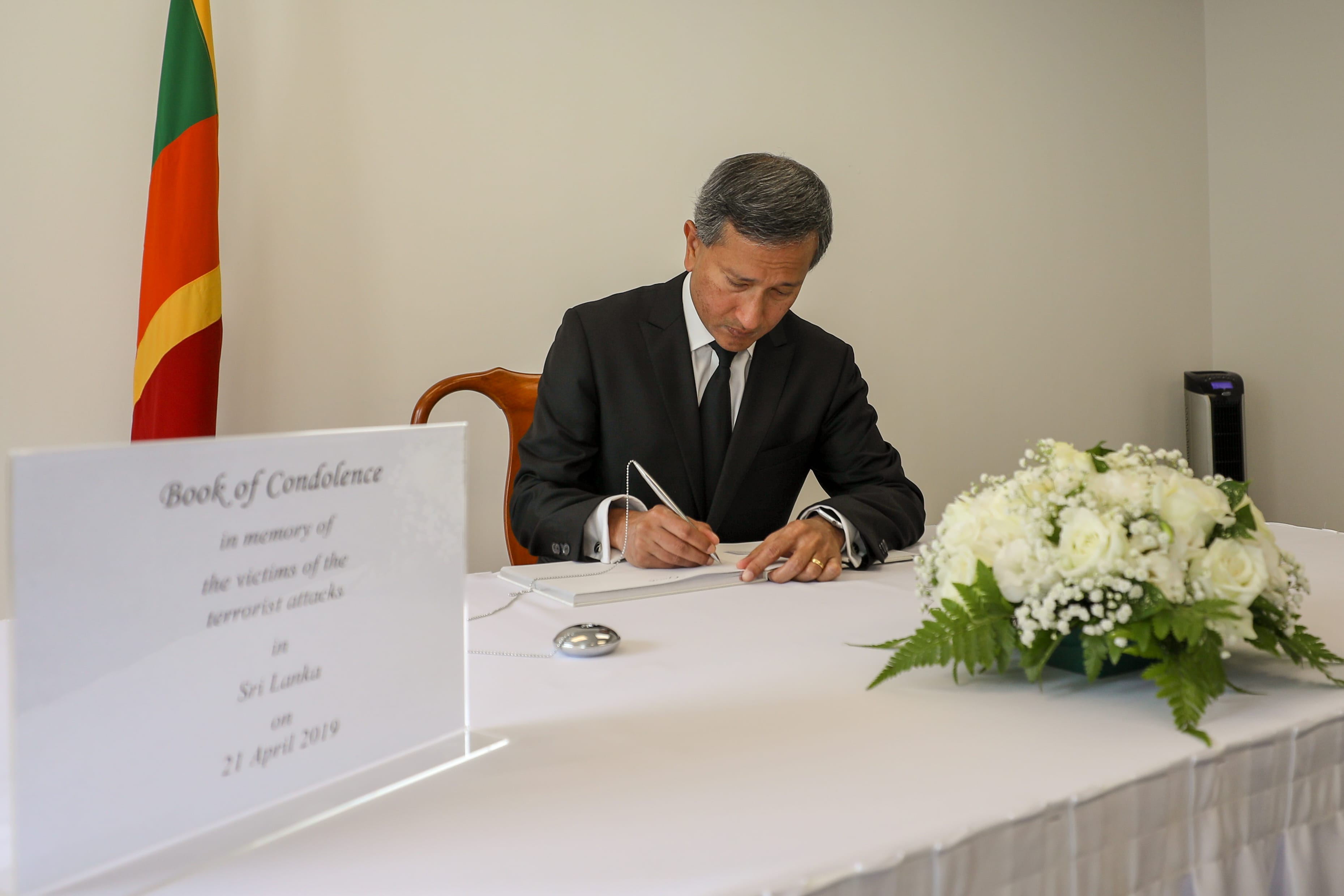 Man in suit signing a Book of Condolence next to the Sri Lankan flag and floral arrangement.