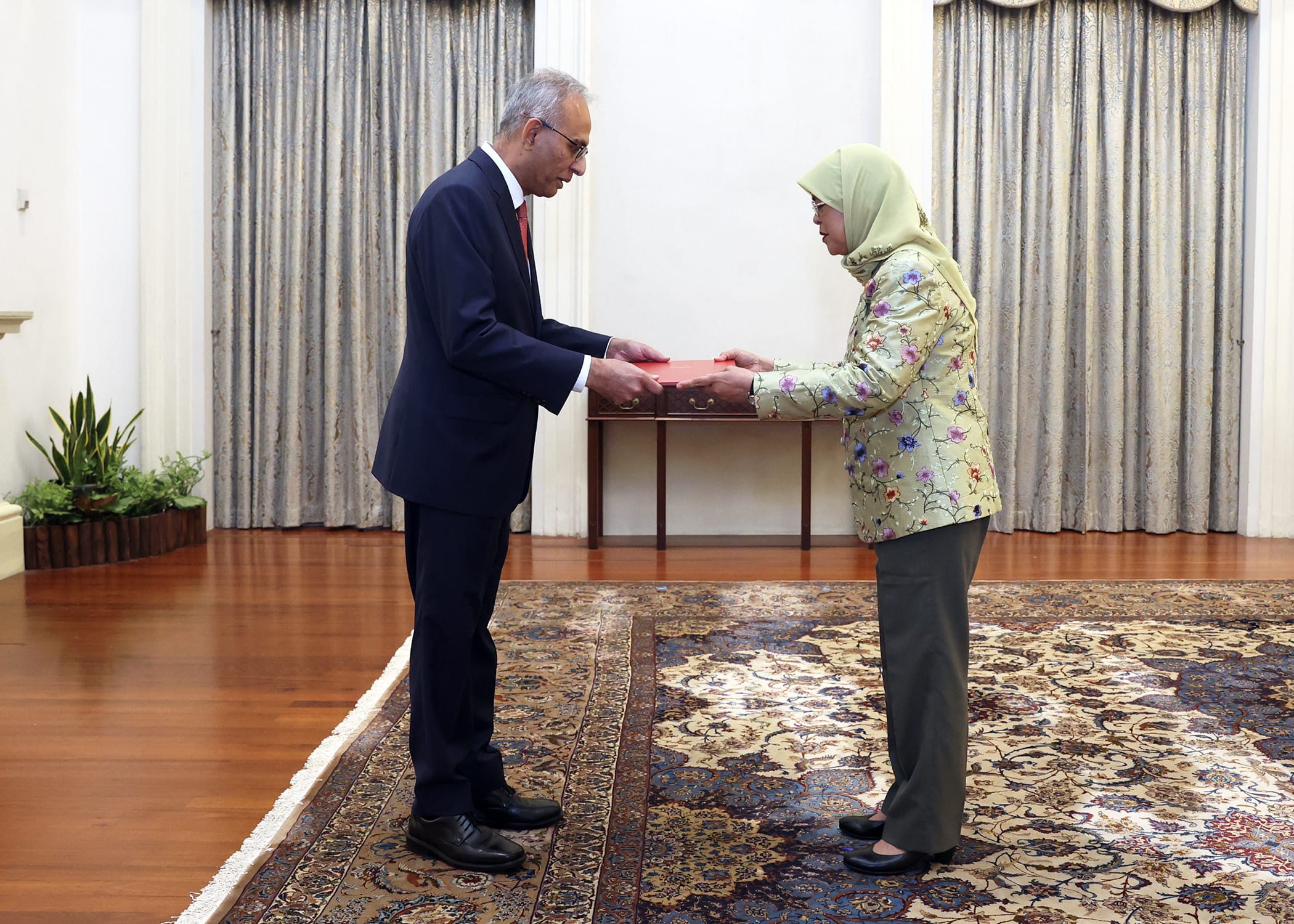 Man in suit hands red box to woman in headscarf and floral jacket.