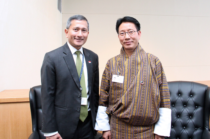 Two men pose indoors; one in suit with tie, other in traditional Bhutanese attire.
