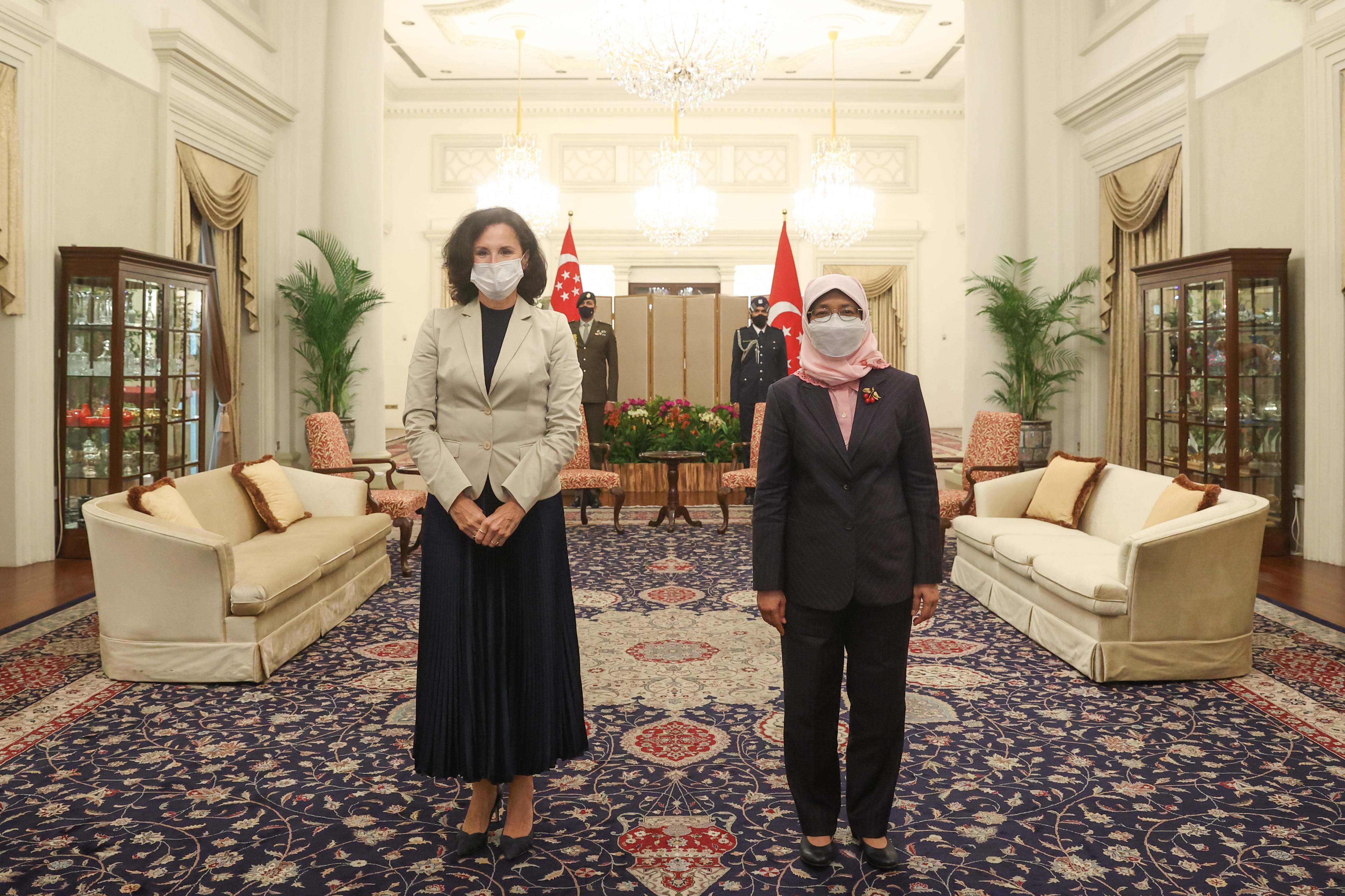 Two women wearing face masks stand in a formal room with flags and guards behind them.