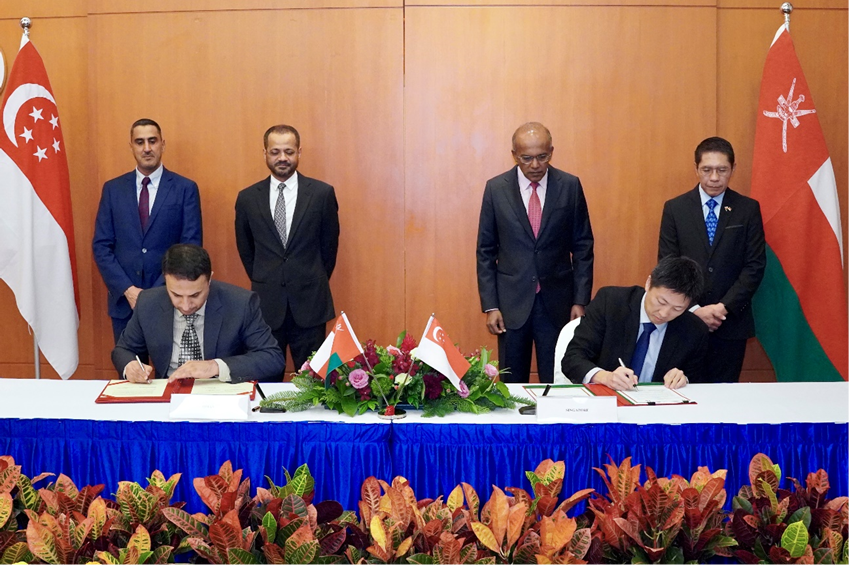 Two people sign agreements, flanked by flags of Singapore and Oman, with observers standing behind.