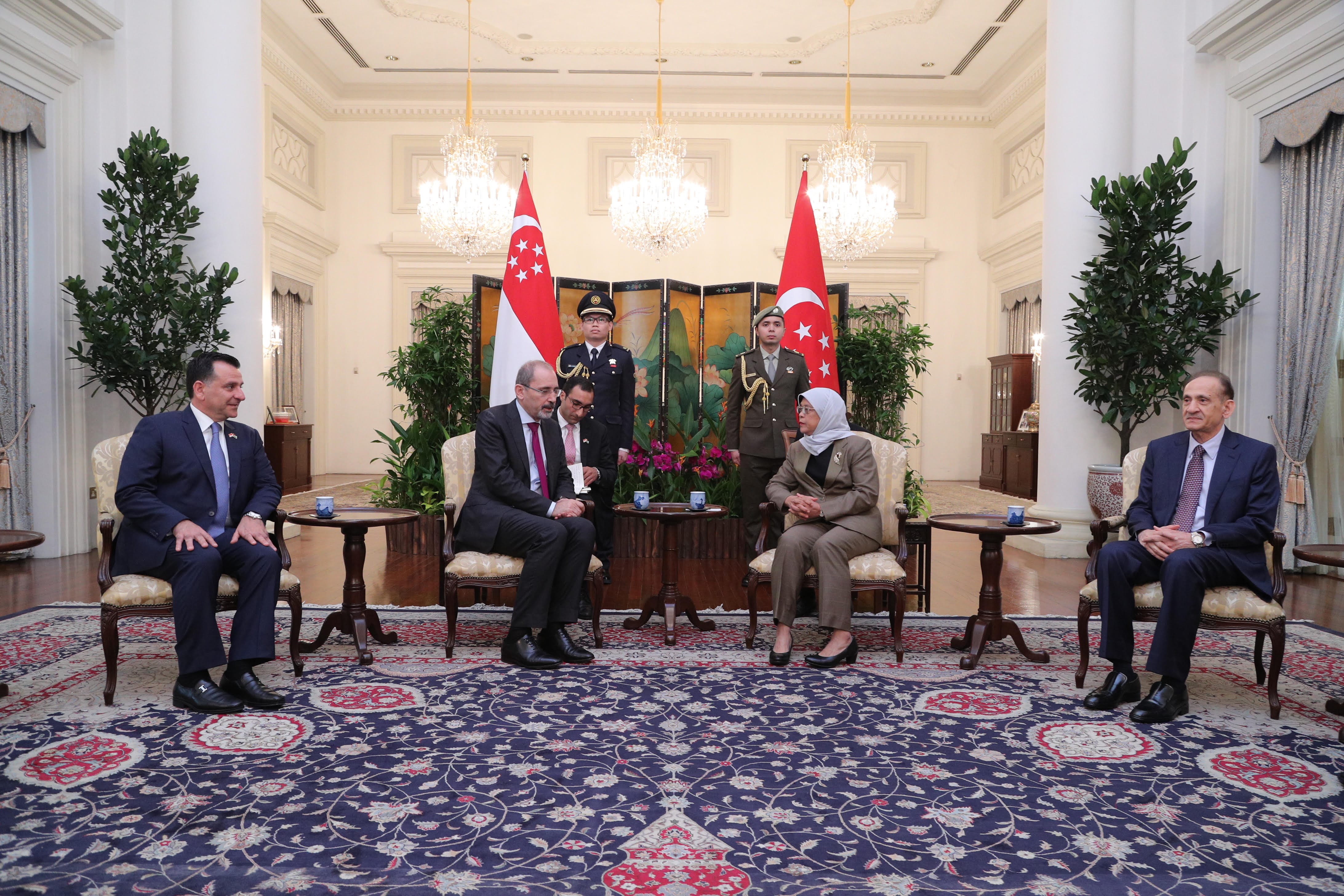 Three men in a room, Kuwaiti flag visible; framed portraits in background.