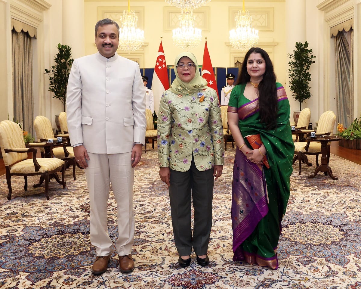 Three people stand indoors before Singapore flags; woman in headscarf wears floral jacket.