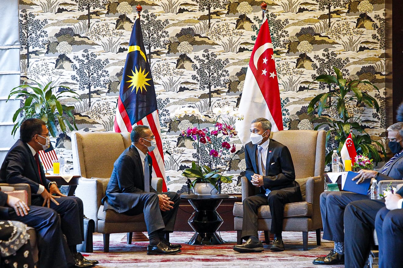 Men in suits and masks sit near Malaysia and Singapore flags in a patterned room.