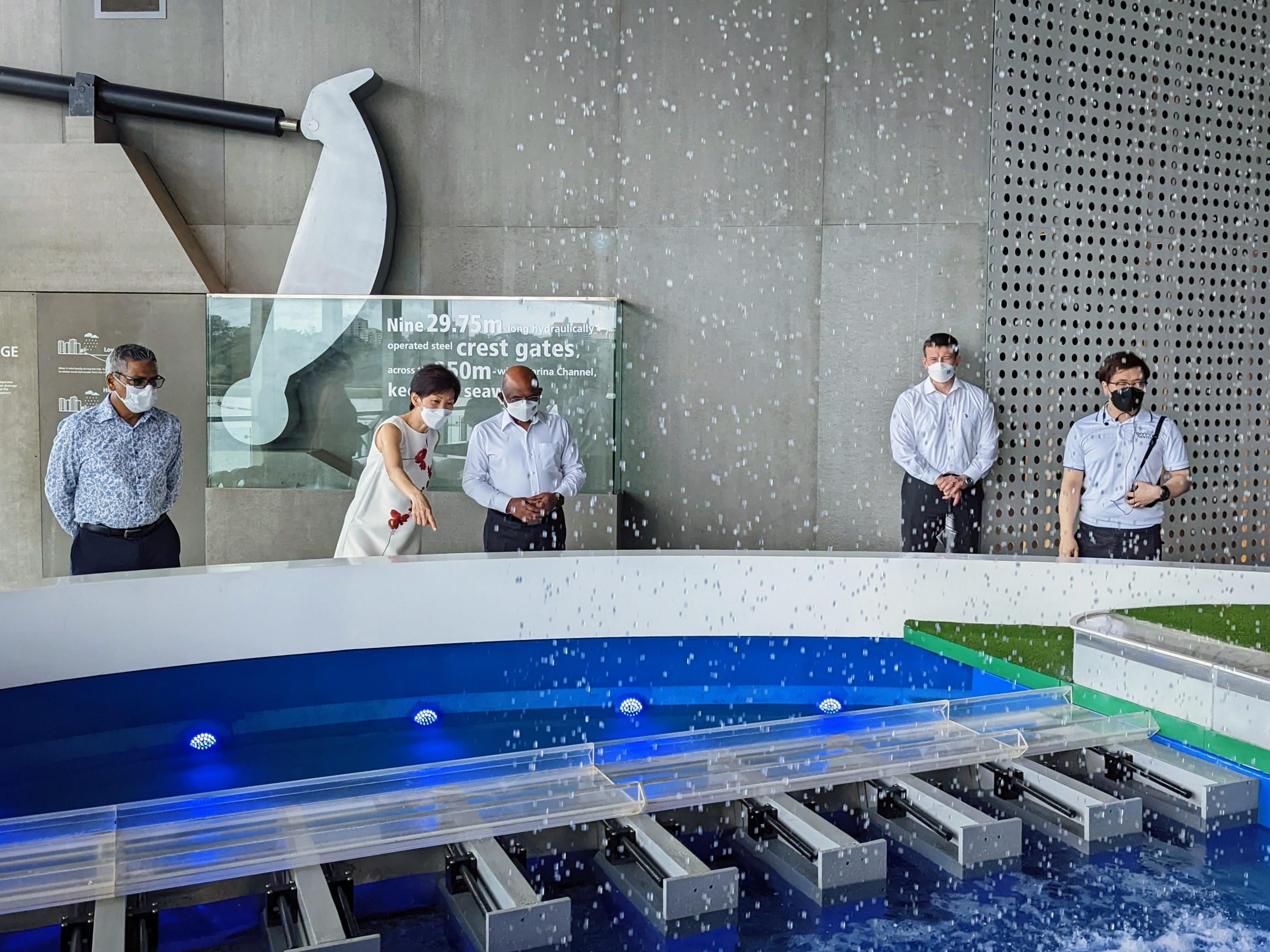 Five people in masks view a water control model with a falling water feature indoors.