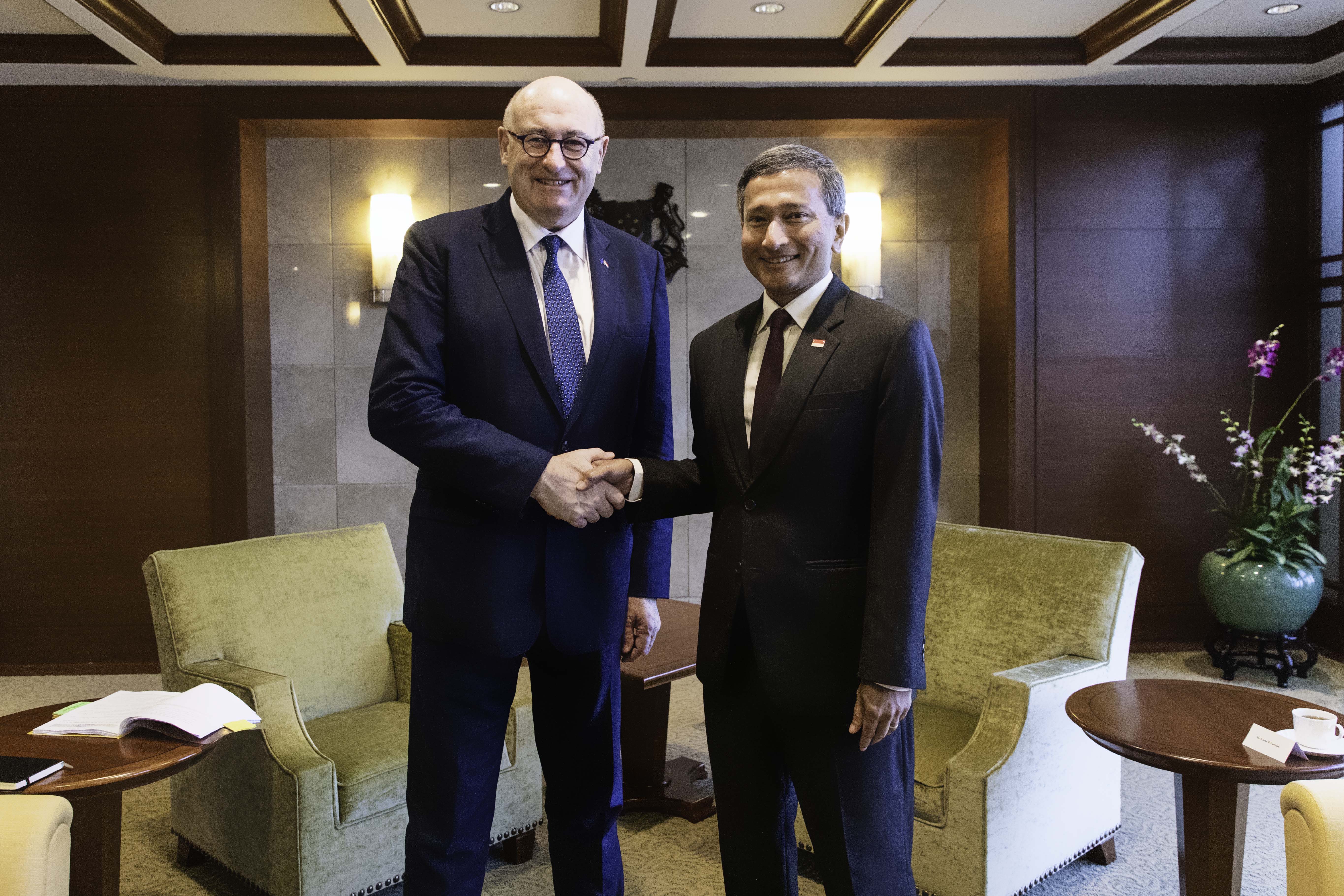 Two men in suits shaking hands in a lounge area with chairs and tables.