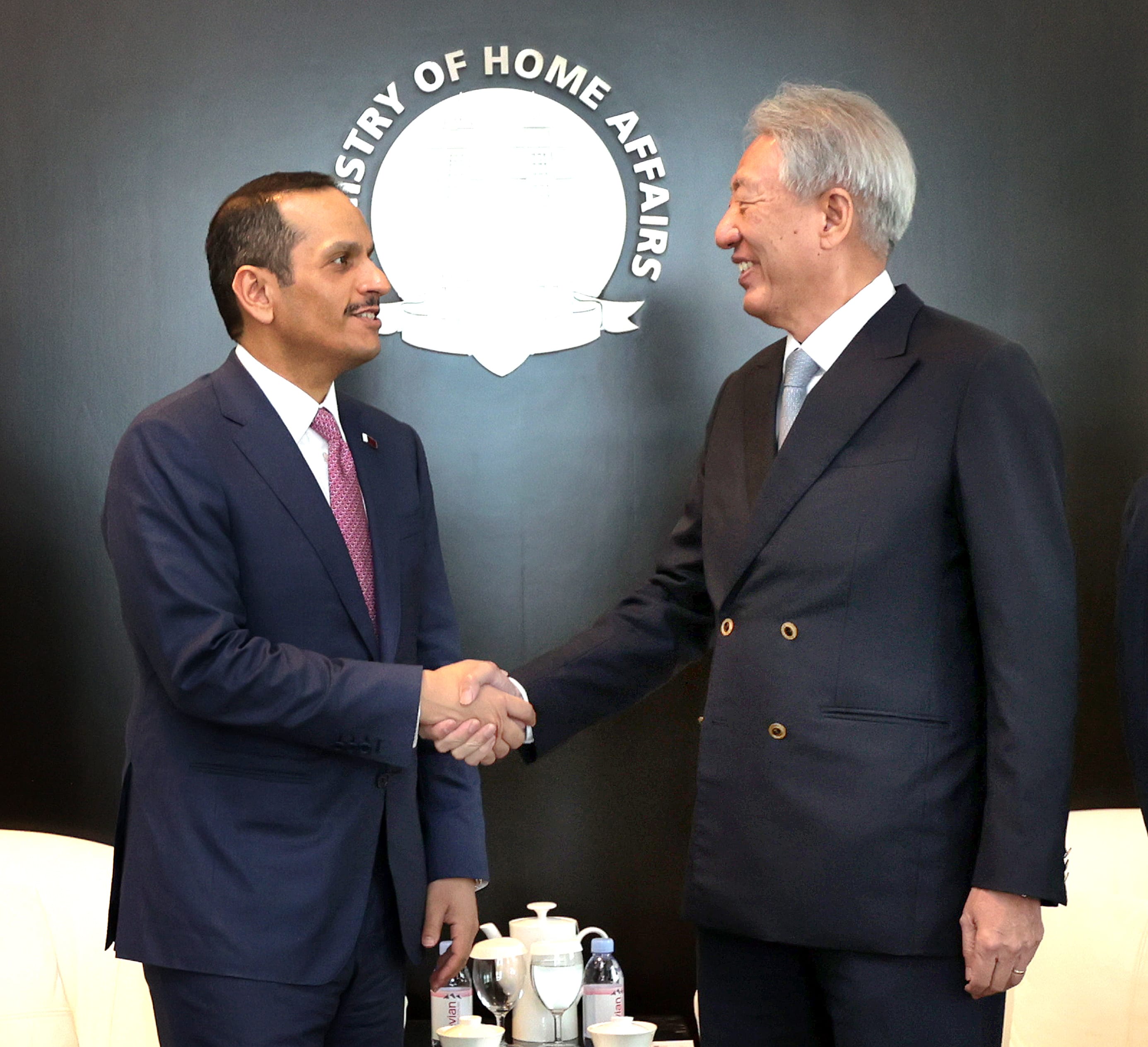 Two men in suits shake hands against a "Ministry of Home Affairs" background.