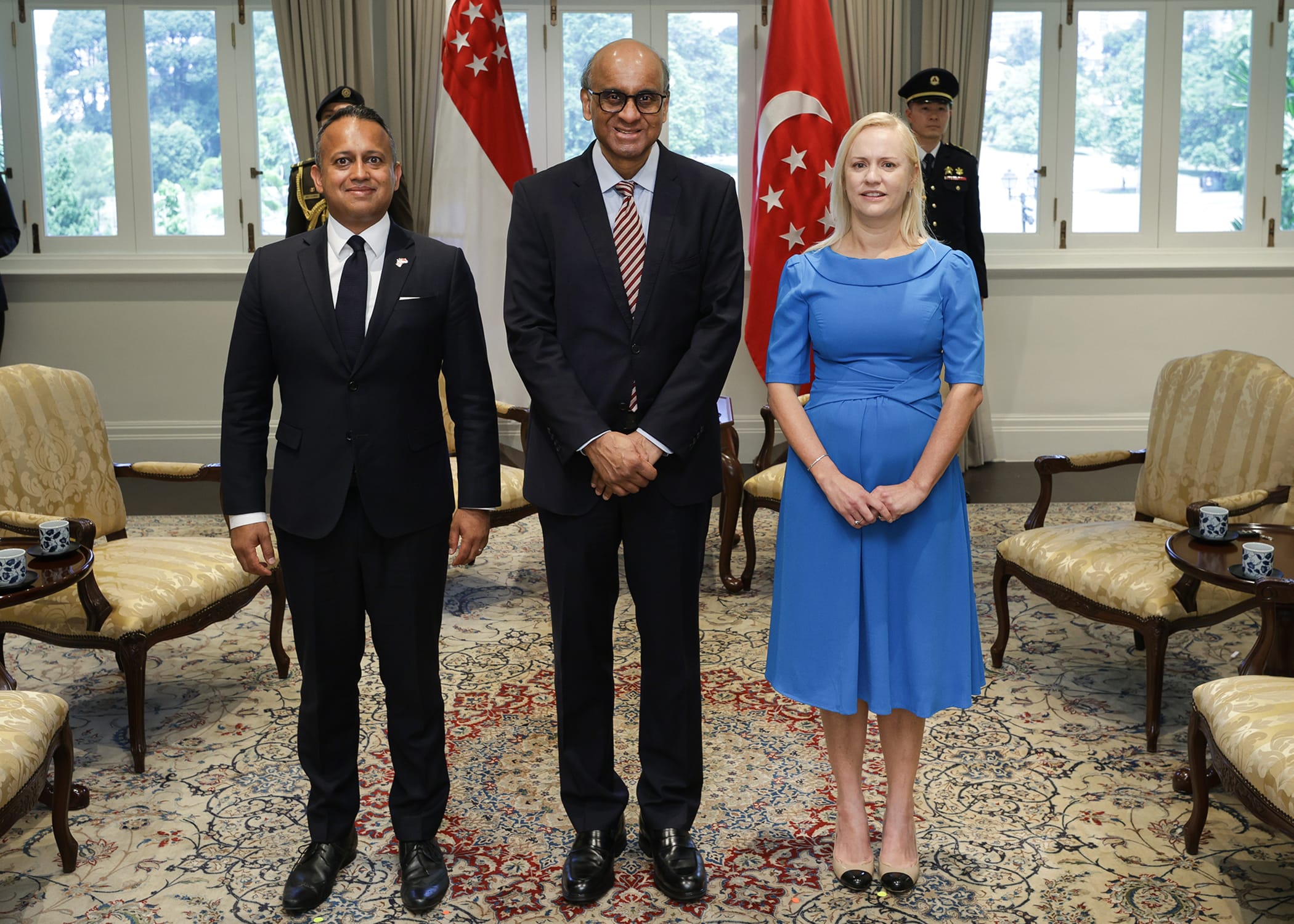 Three people stand before Singapore flags, two men in suits, woman in blue dress.