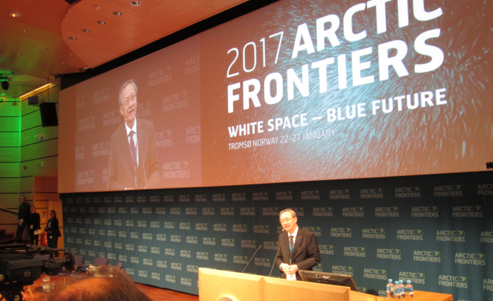 Man at podium at "Arctic Frontiers" event; large screen displays speaker and event title.