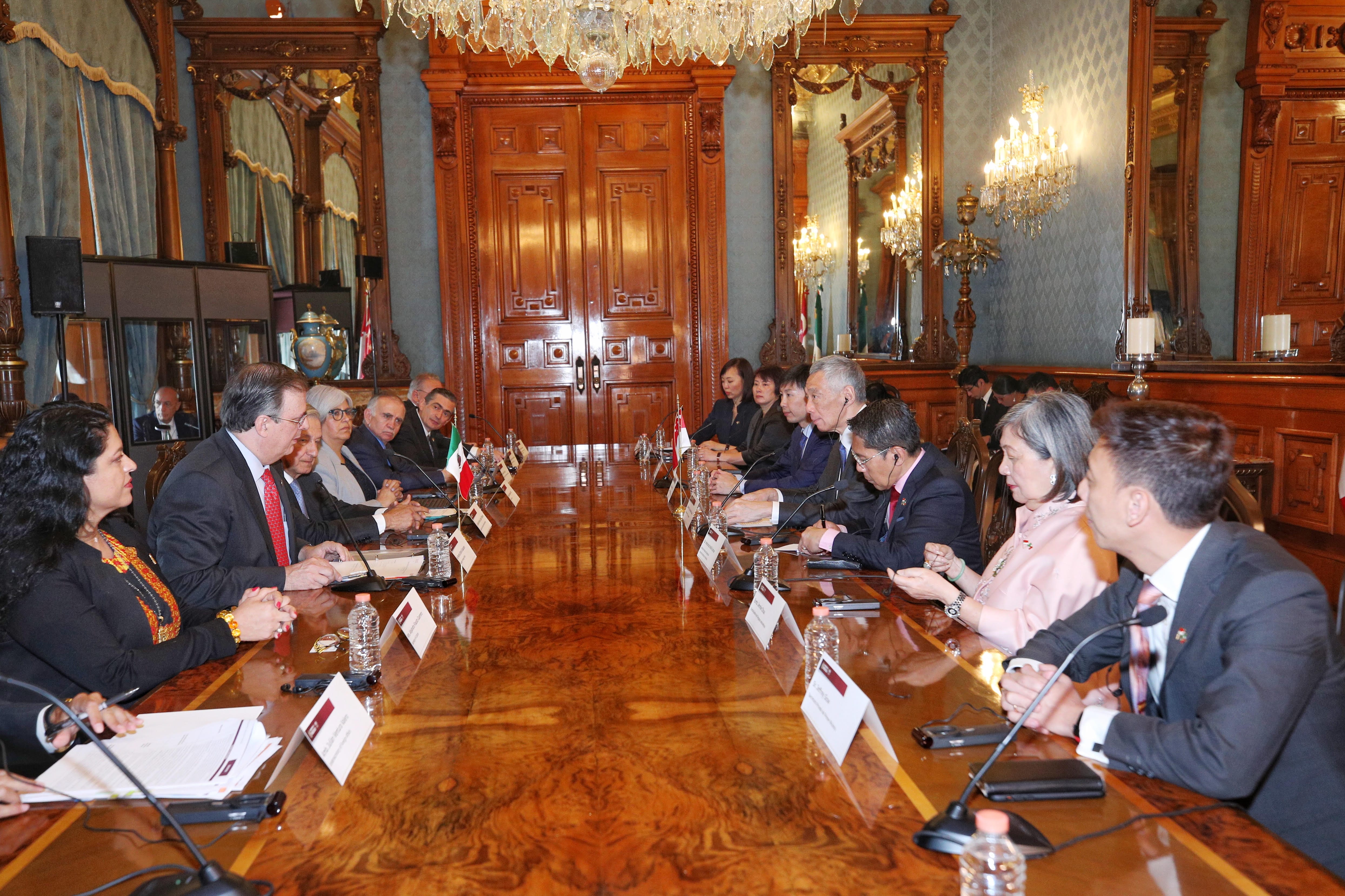 Two men hold a gift-wrapped box in a room with framed portraits and Kuwaiti flag.