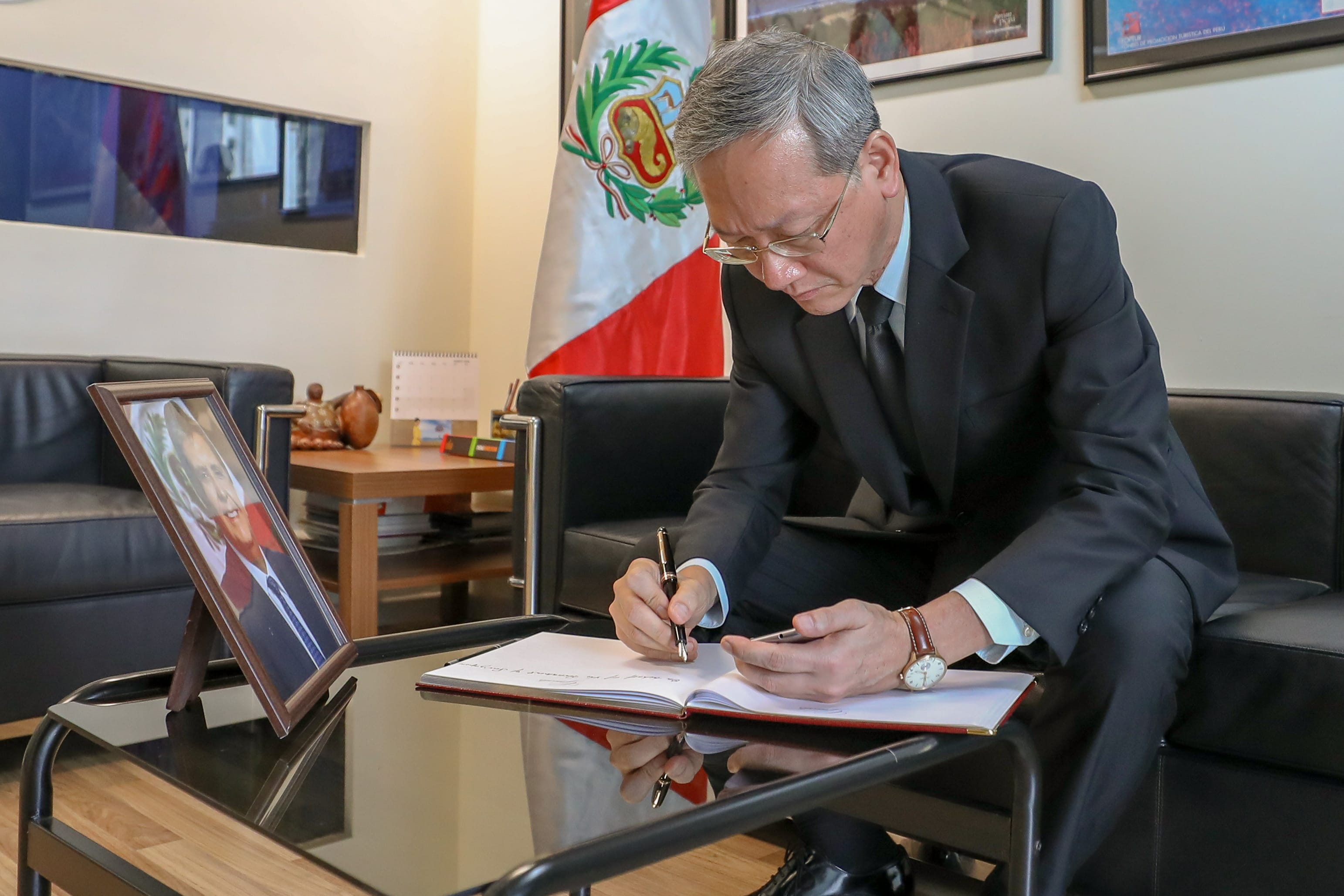 Man signs book with Peruvian flag. Framed portrait visible on table.