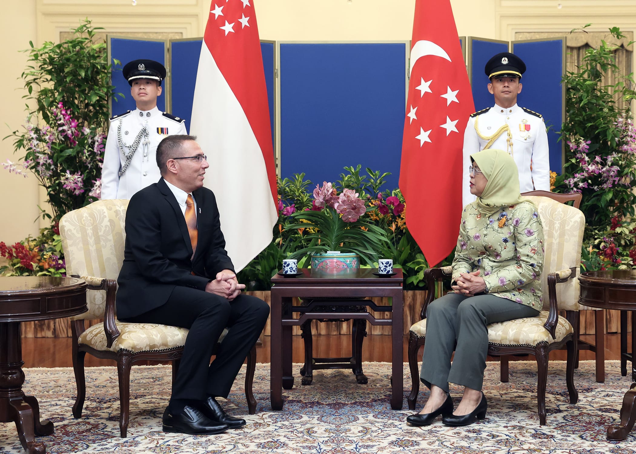 Two people seated, facing each other, flanked by Singapore flags and guards in ceremonial dress.