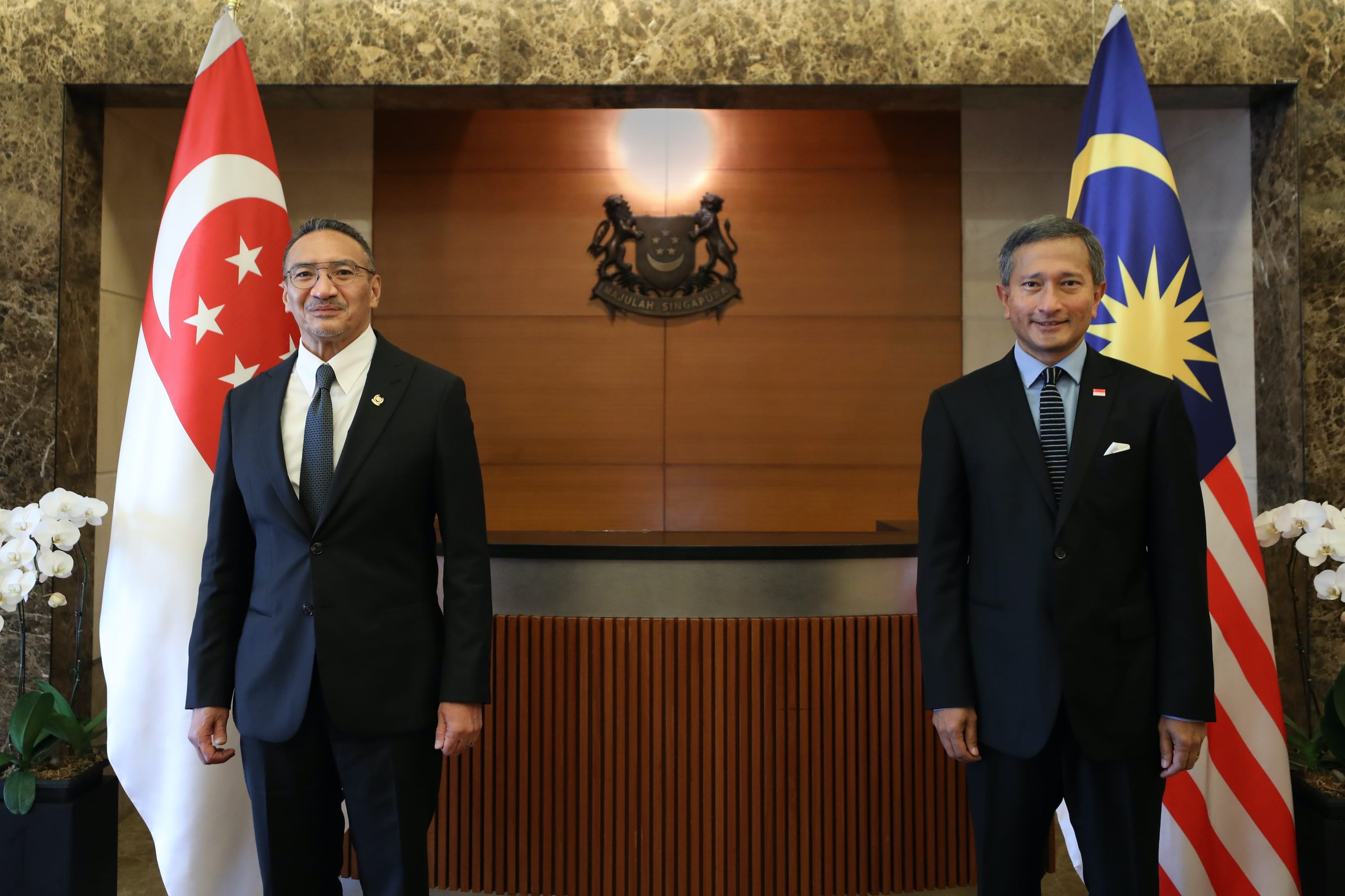 Two men in suits stand beside Singapore and Malaysia flags.