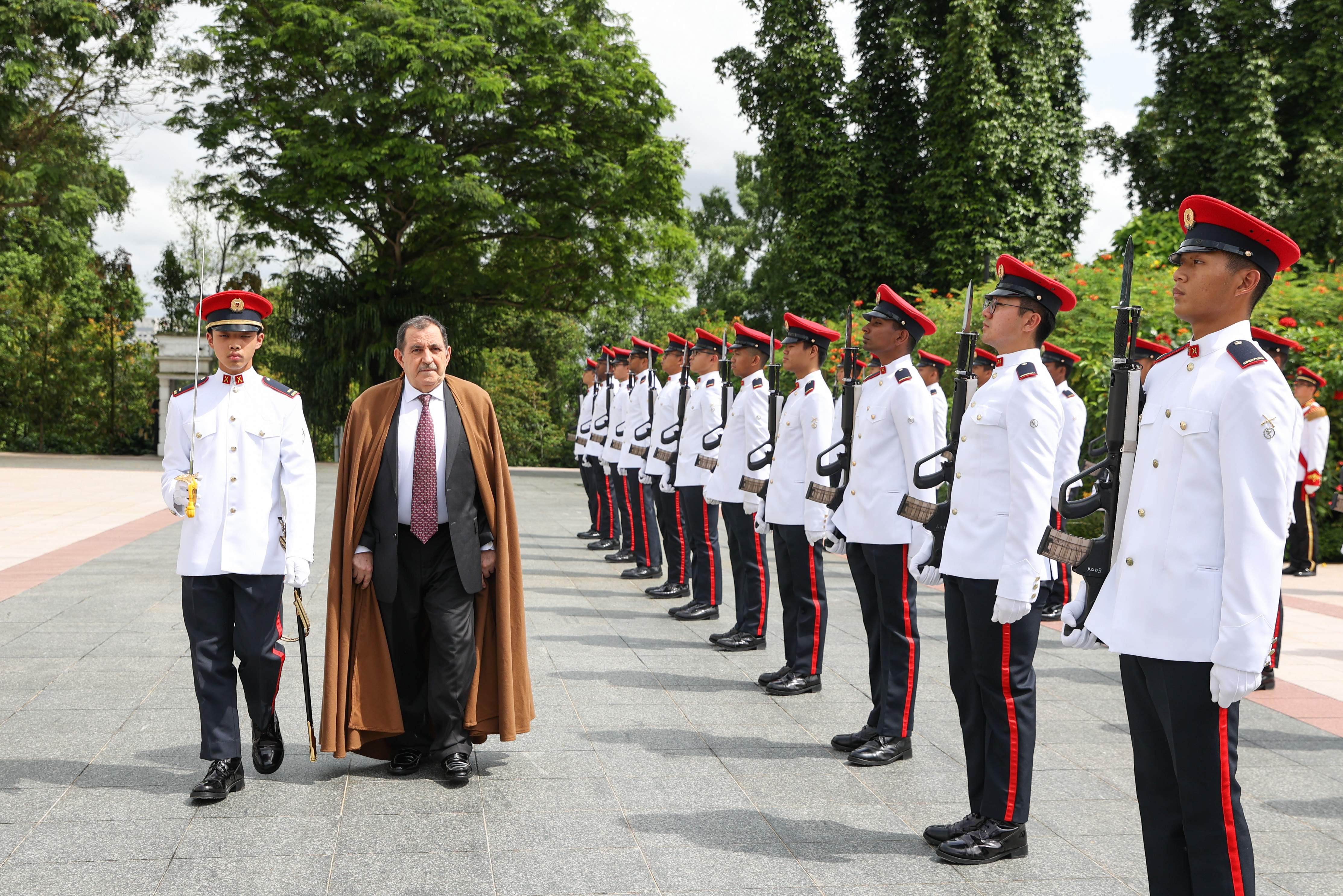 Line of soldiers with rifles at attention; man in suit/cape walking nearby.