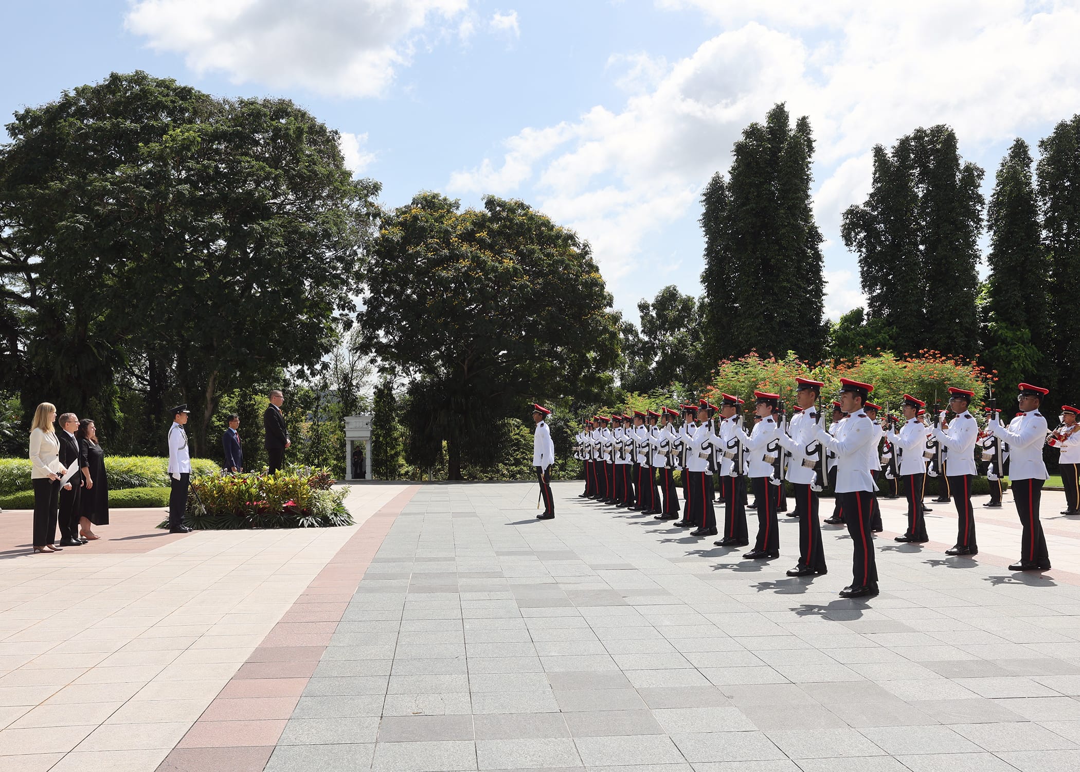 Honor guard in white uniforms and red hats stand at attention before officials outdoors.