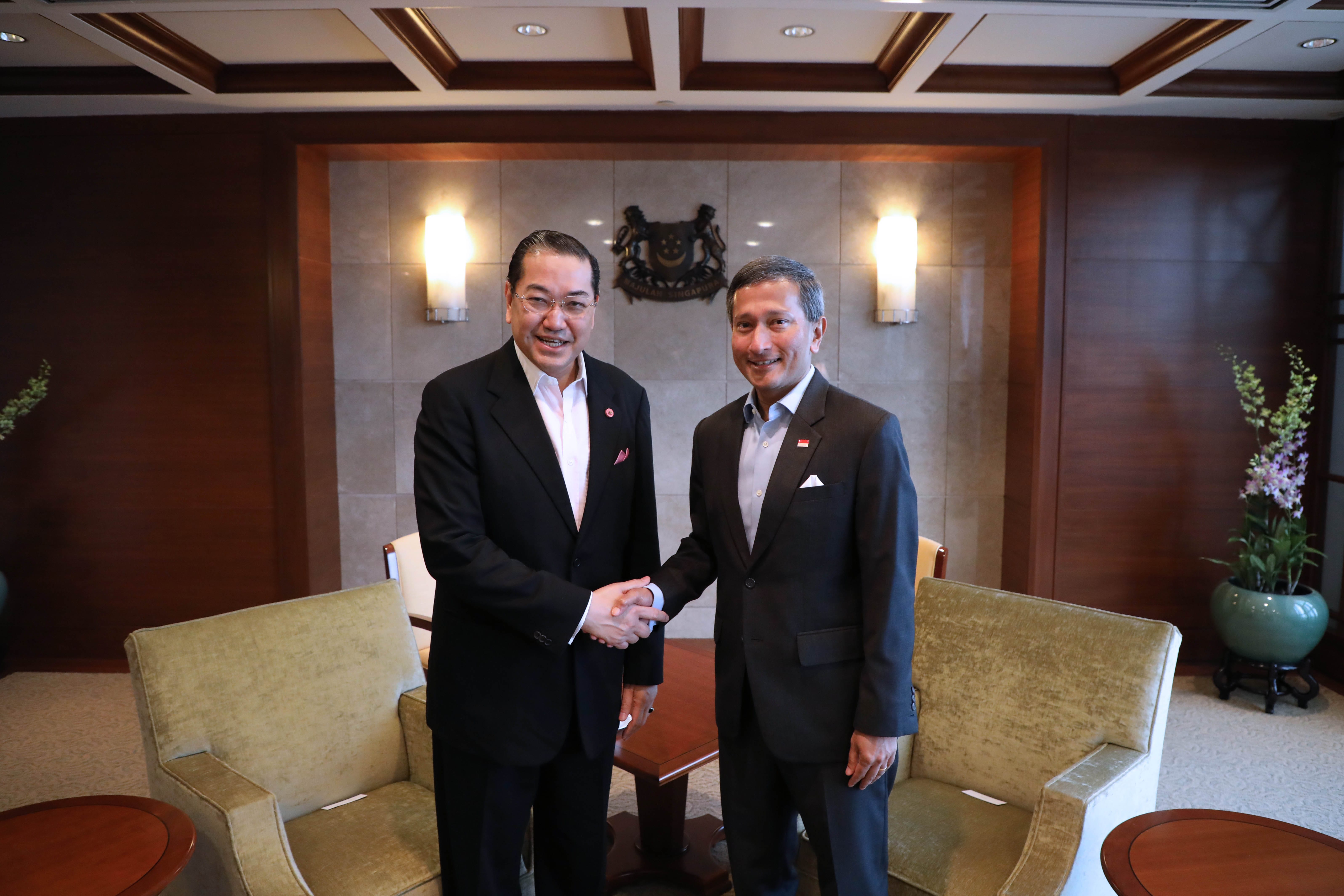 Two men in suits shake hands in front of a wall with the Singapore coat of arms.