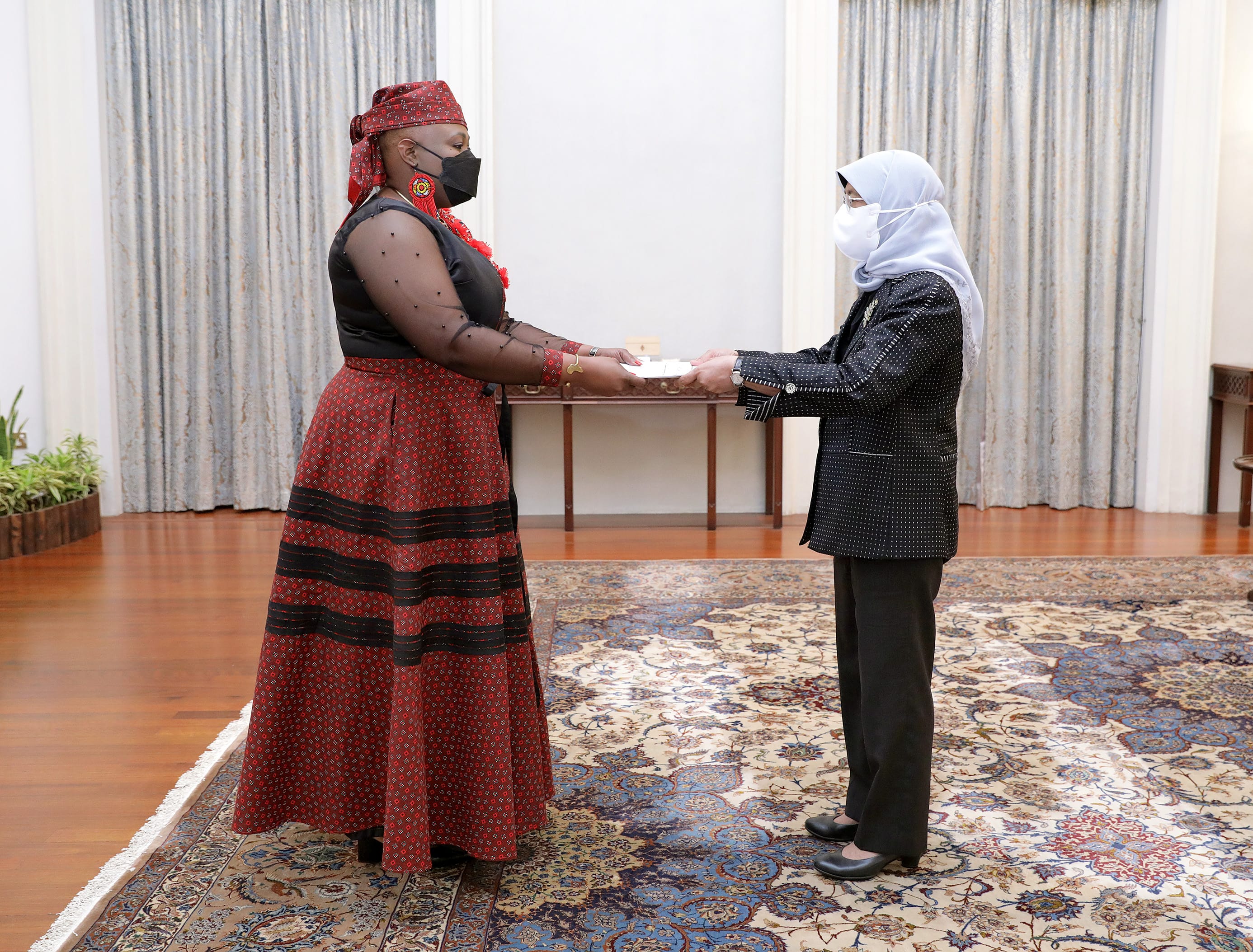 Two people in face masks exchanging papers in a room with patterned carpet.