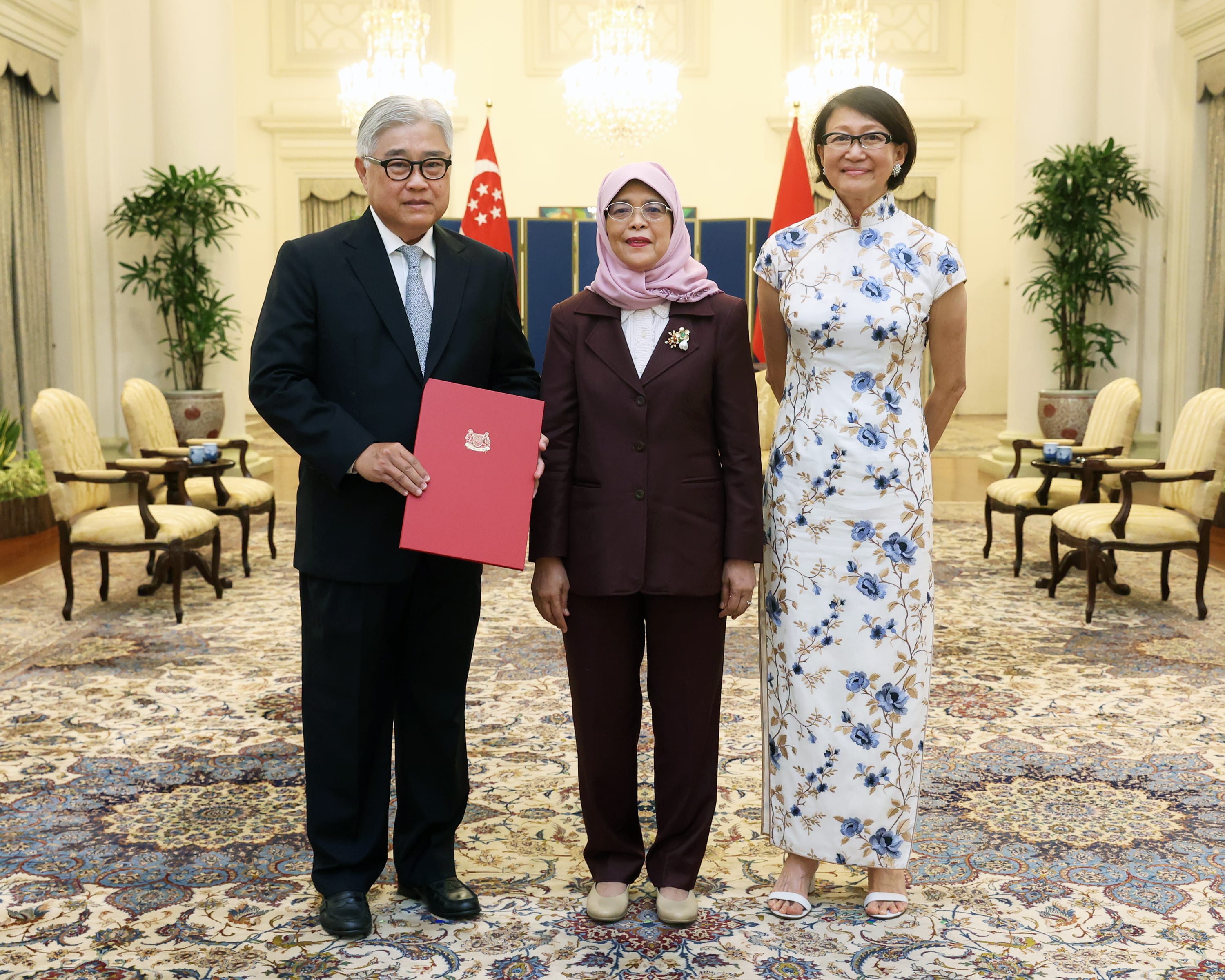 Three people in formal attire pose in front of Singapore flags, holding a red folder.