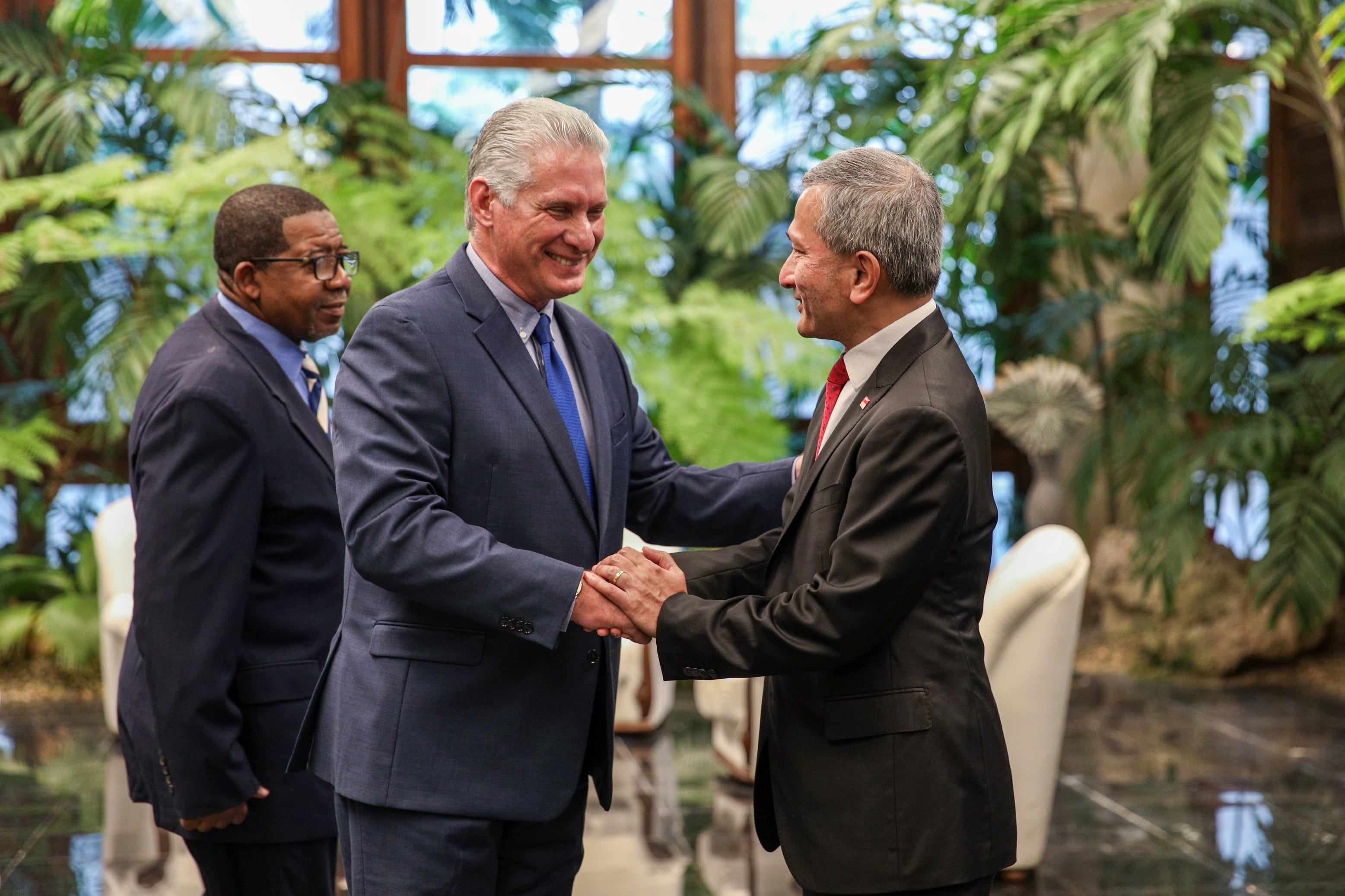 Three men in suits, two shaking hands. Lush green foliage in background.