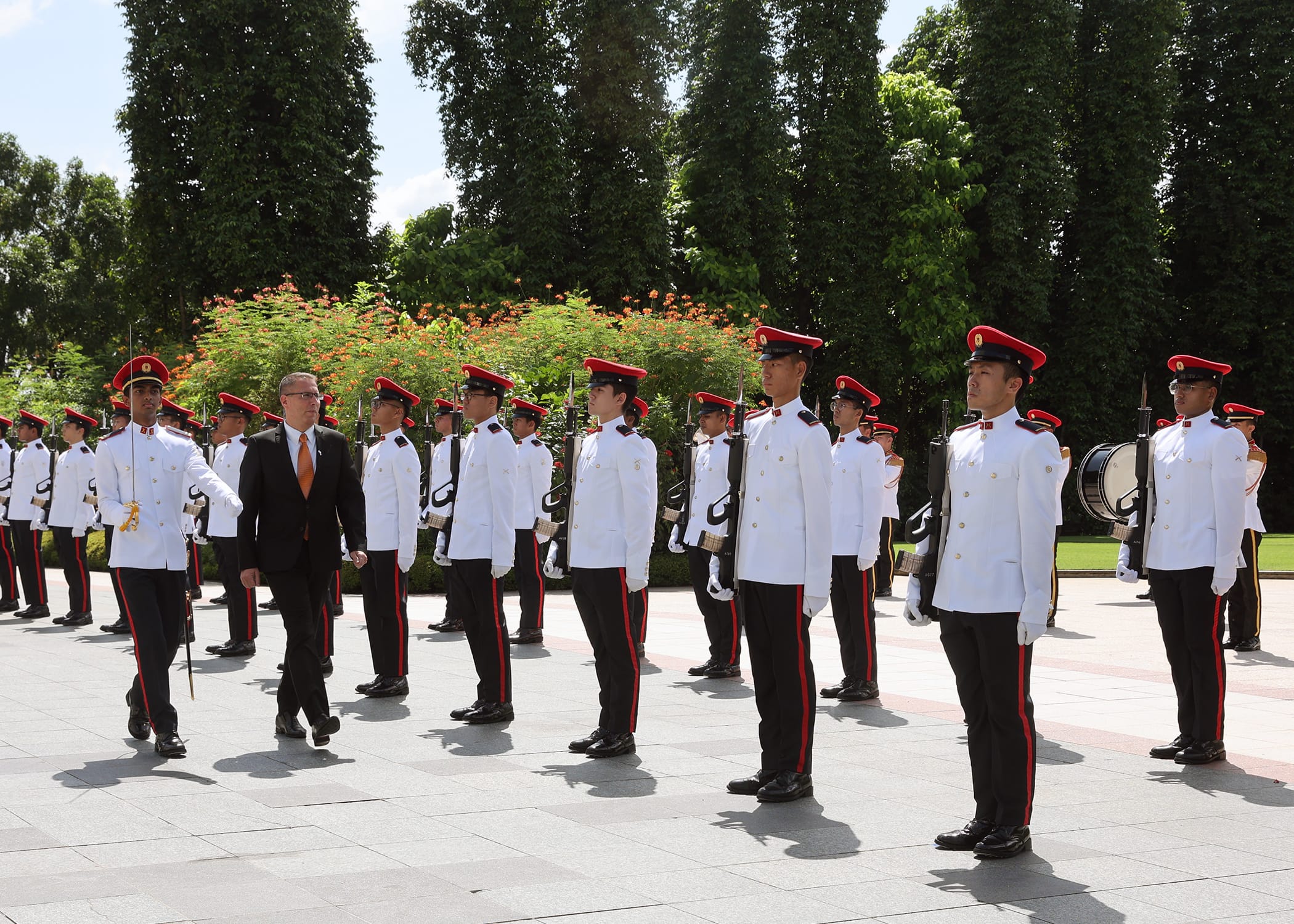 Military honor guard in white uniforms and red hats, with a man in a suit, outdoors.