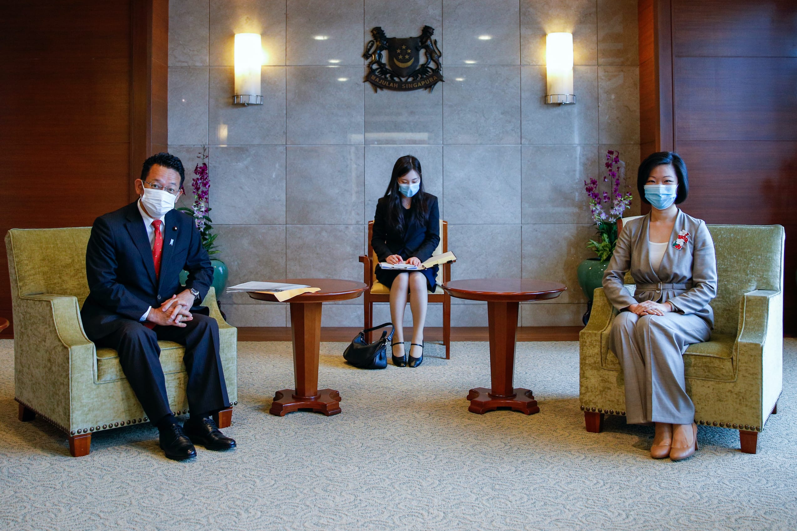 Three masked people seated in armchairs in front of a Singapore crest on a wall.