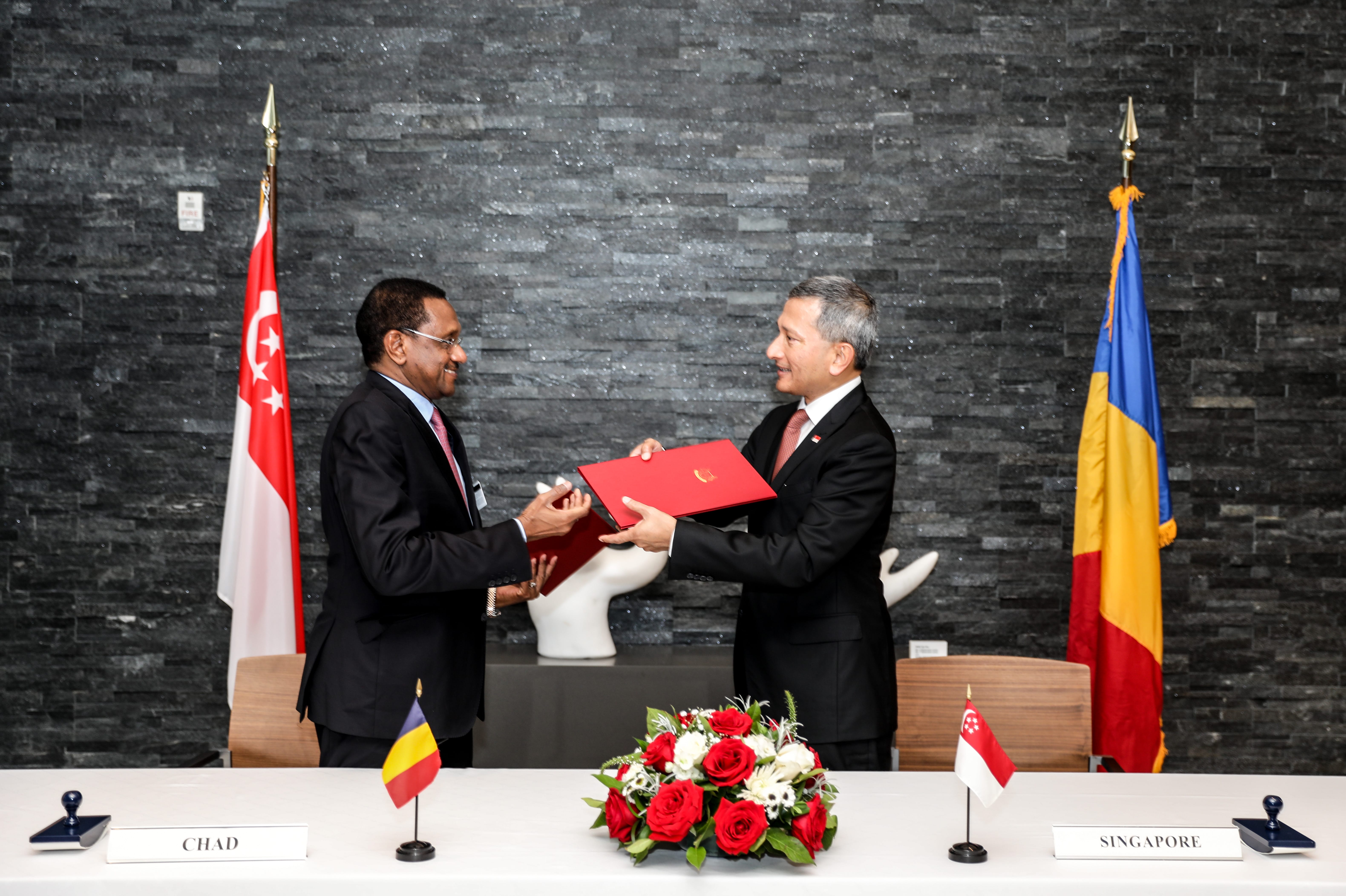 Two men exchanging a red document with Singapore & Chad flags on table.