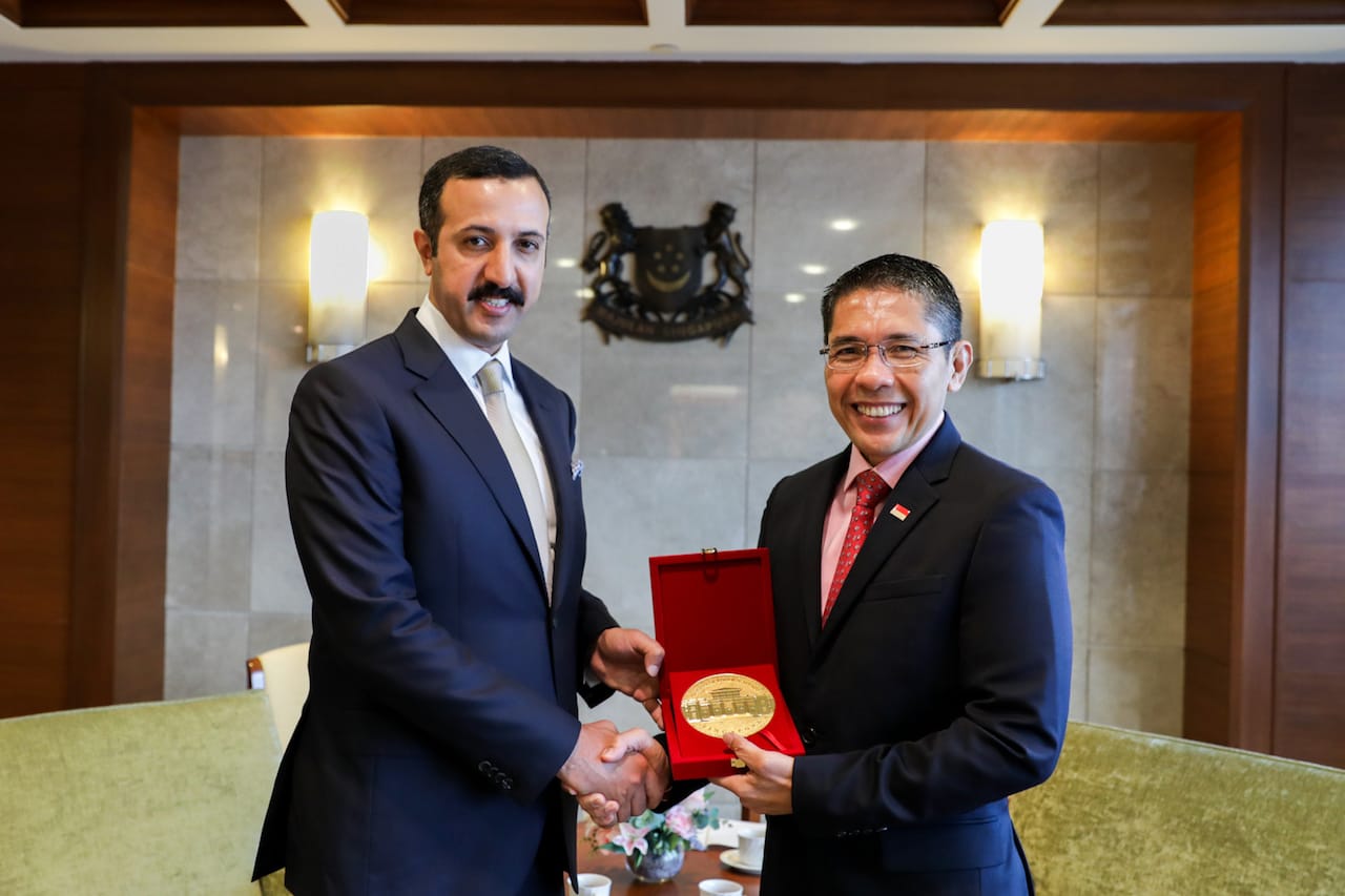Two men in suits shaking hands, one holding a red box with a gold medal. Singapore crest visible.