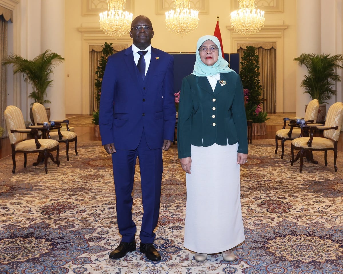 Two people stand indoors, on a patterned carpet. One wears a blue suit; the other, a green jacket.