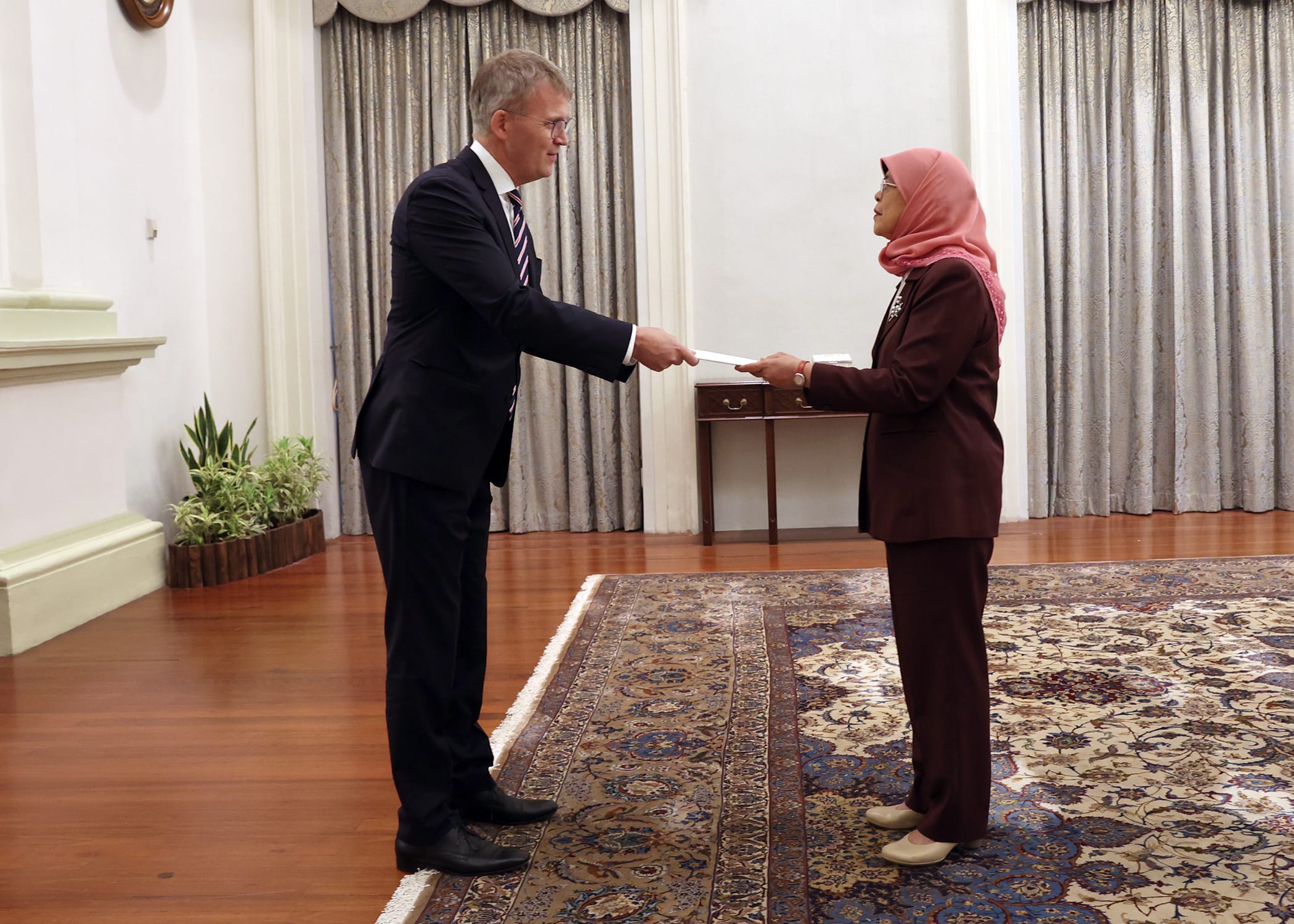 Man in suit presents document to woman in hijab and suit, both indoors on ornate rug.