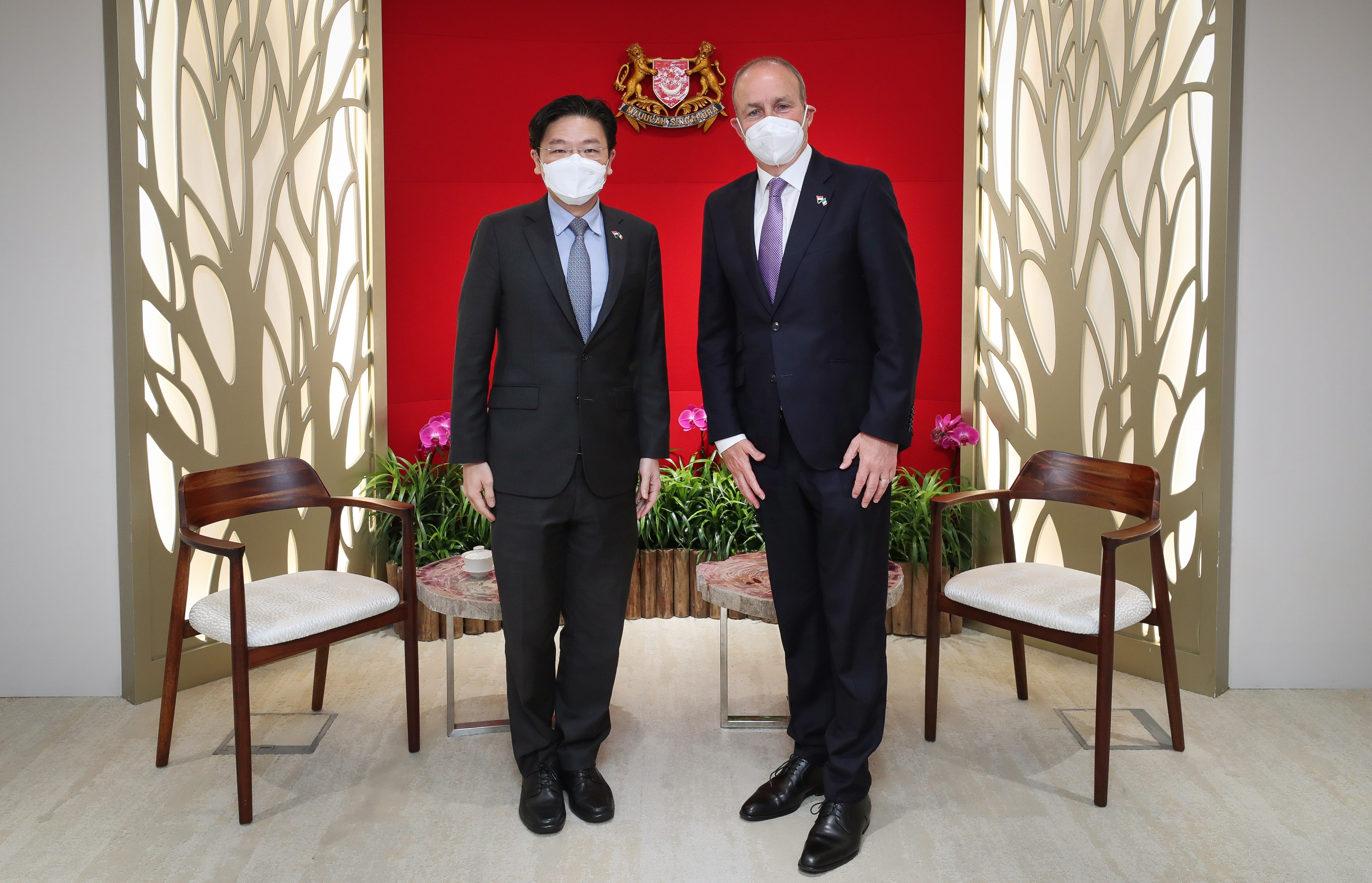 Two men in suits and face masks pose in front of a red wall with Singapore coat of arms.