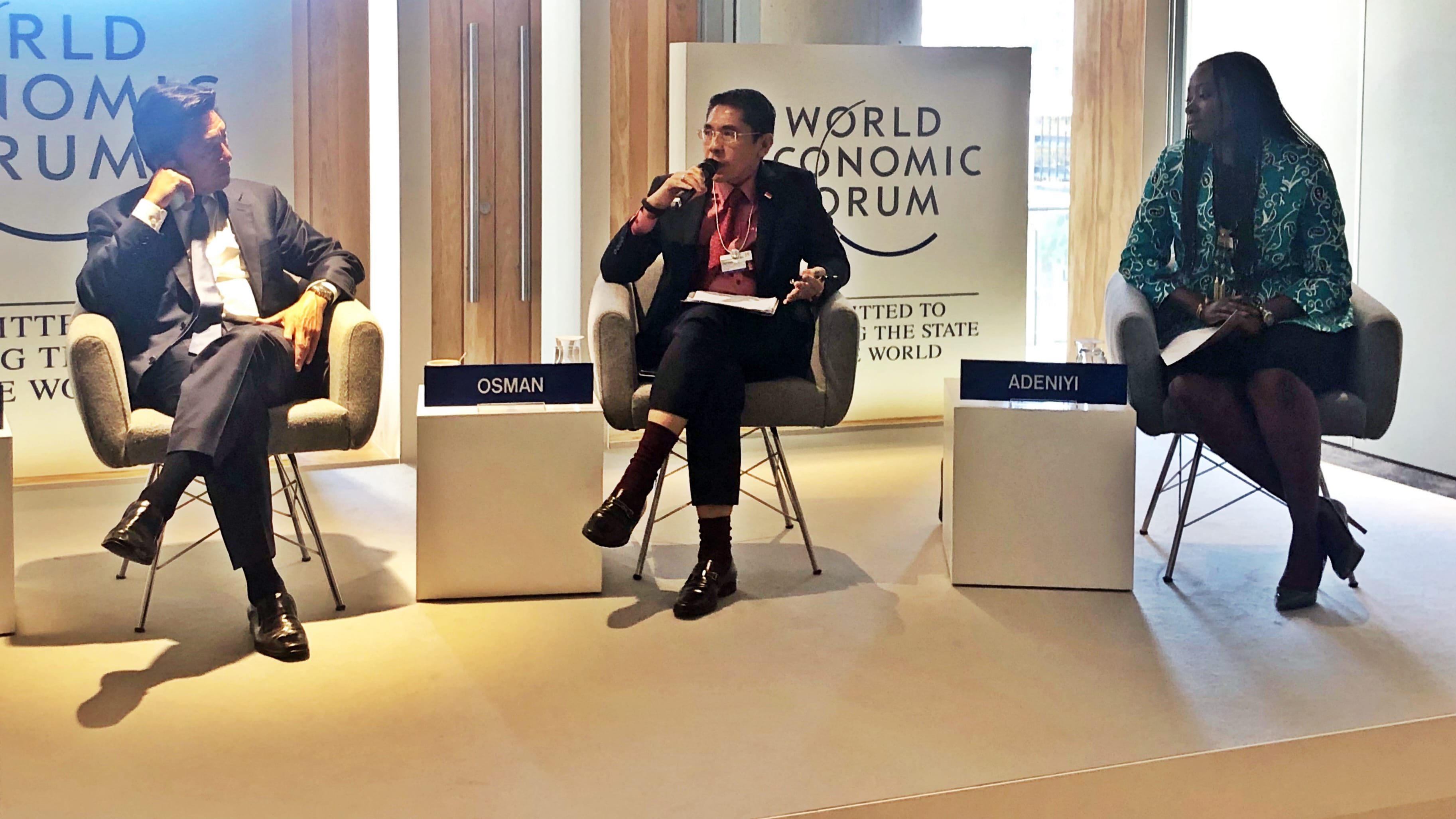 Three men in a room, Kuwaiti flag visible; framed portraits in background.