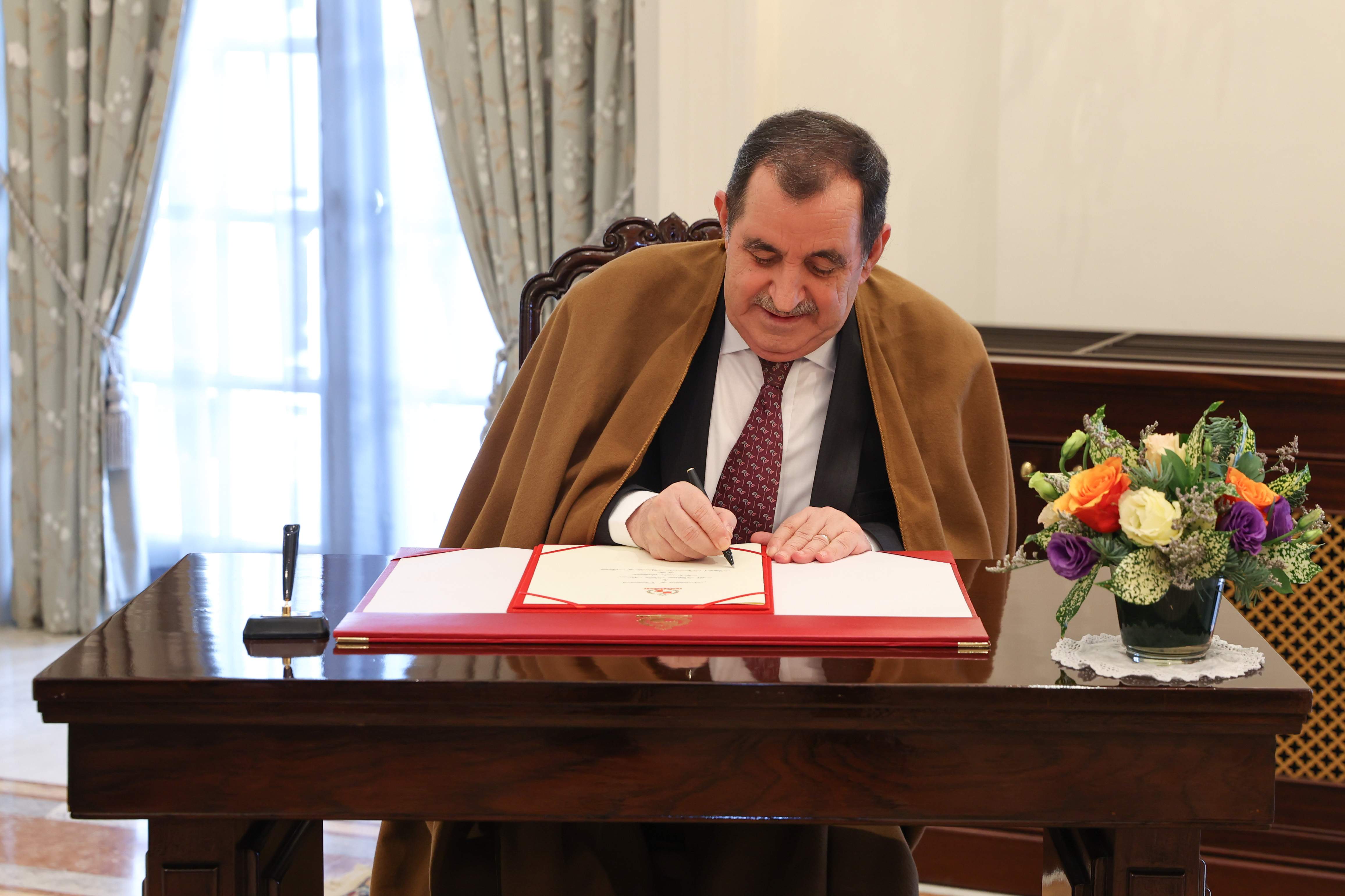 Man in suit and brown robe signing document on a desk. Flowers are beside him.