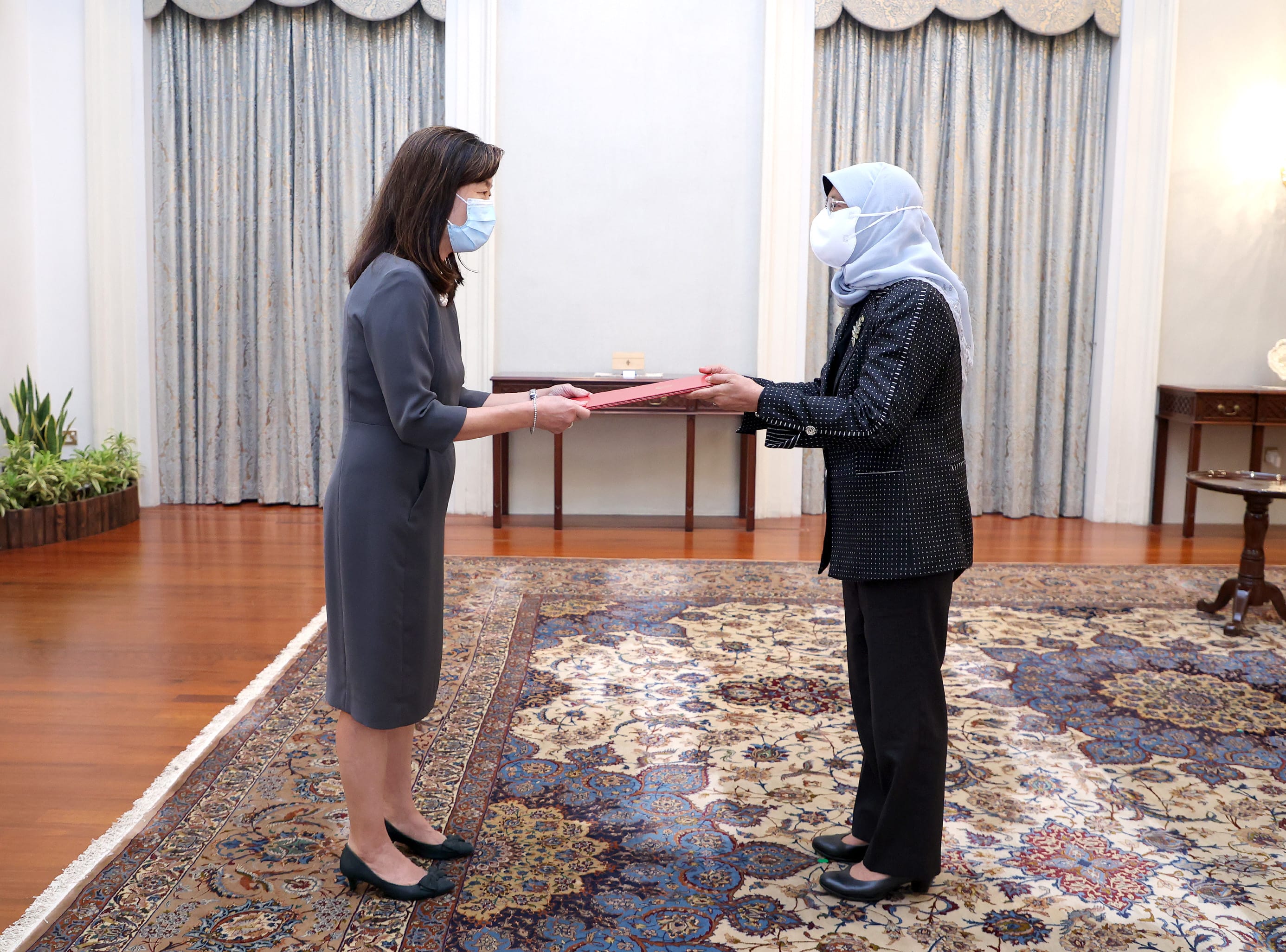 Two masked women exchanging red folders in a room with a patterned rug.