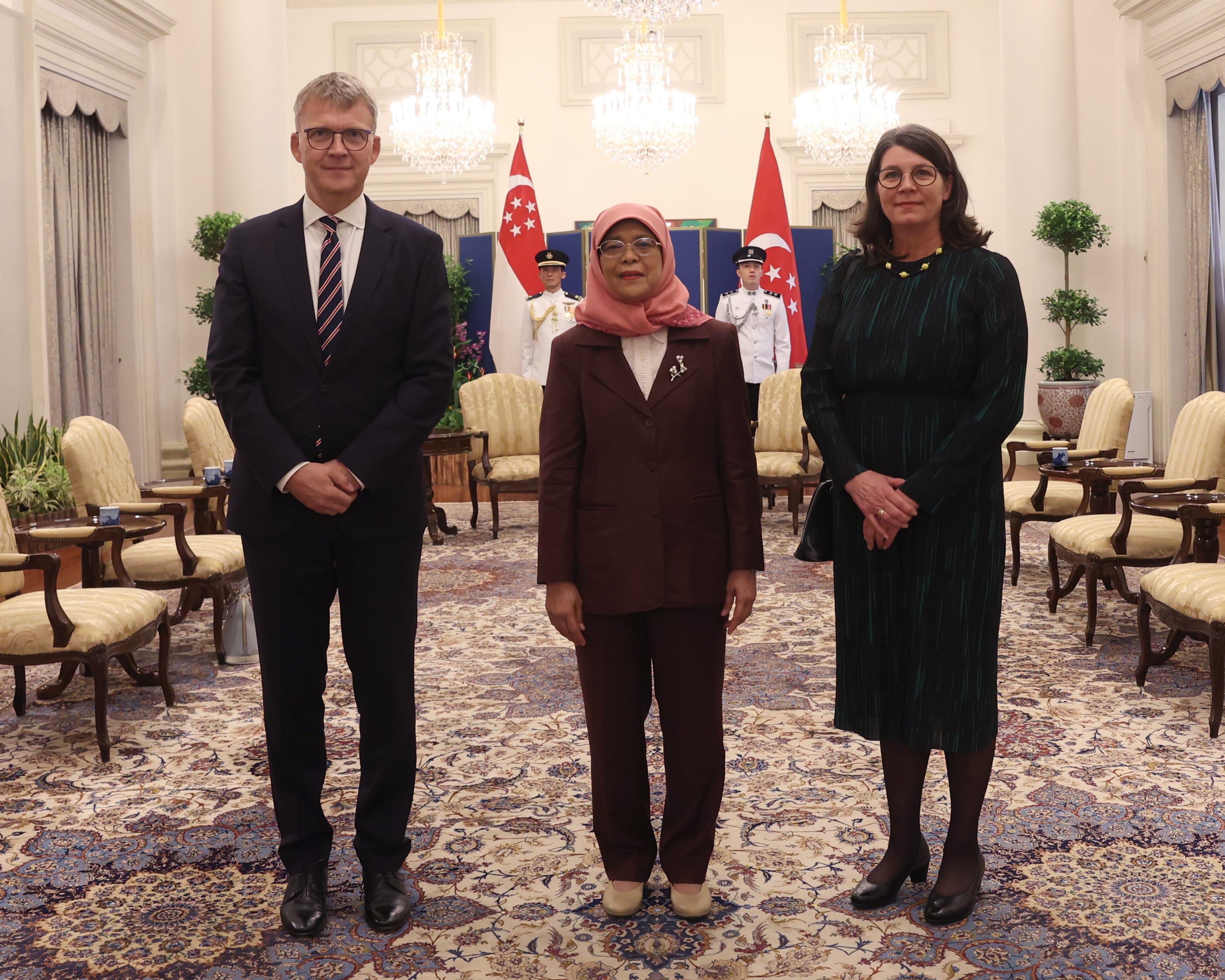Three people stand in a formal setting, Singapore flag behind center figure.