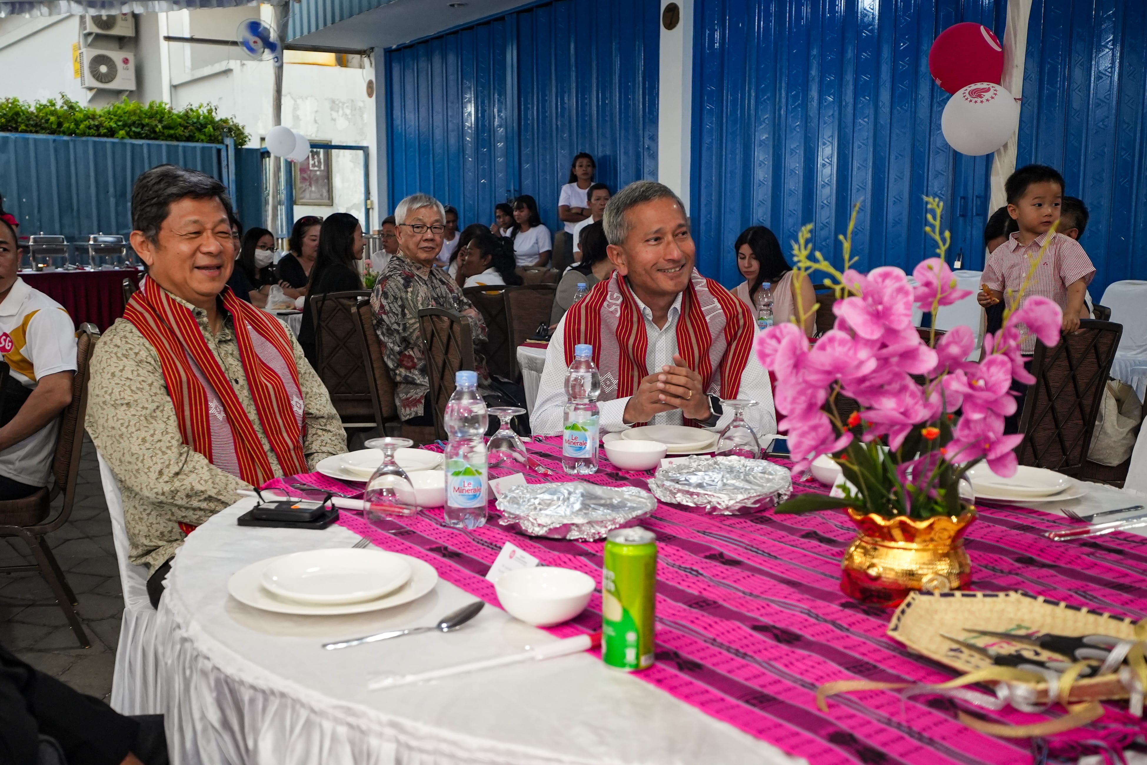 People seated at a table with pink tablecloth, pink flowers, water bottles, and food.