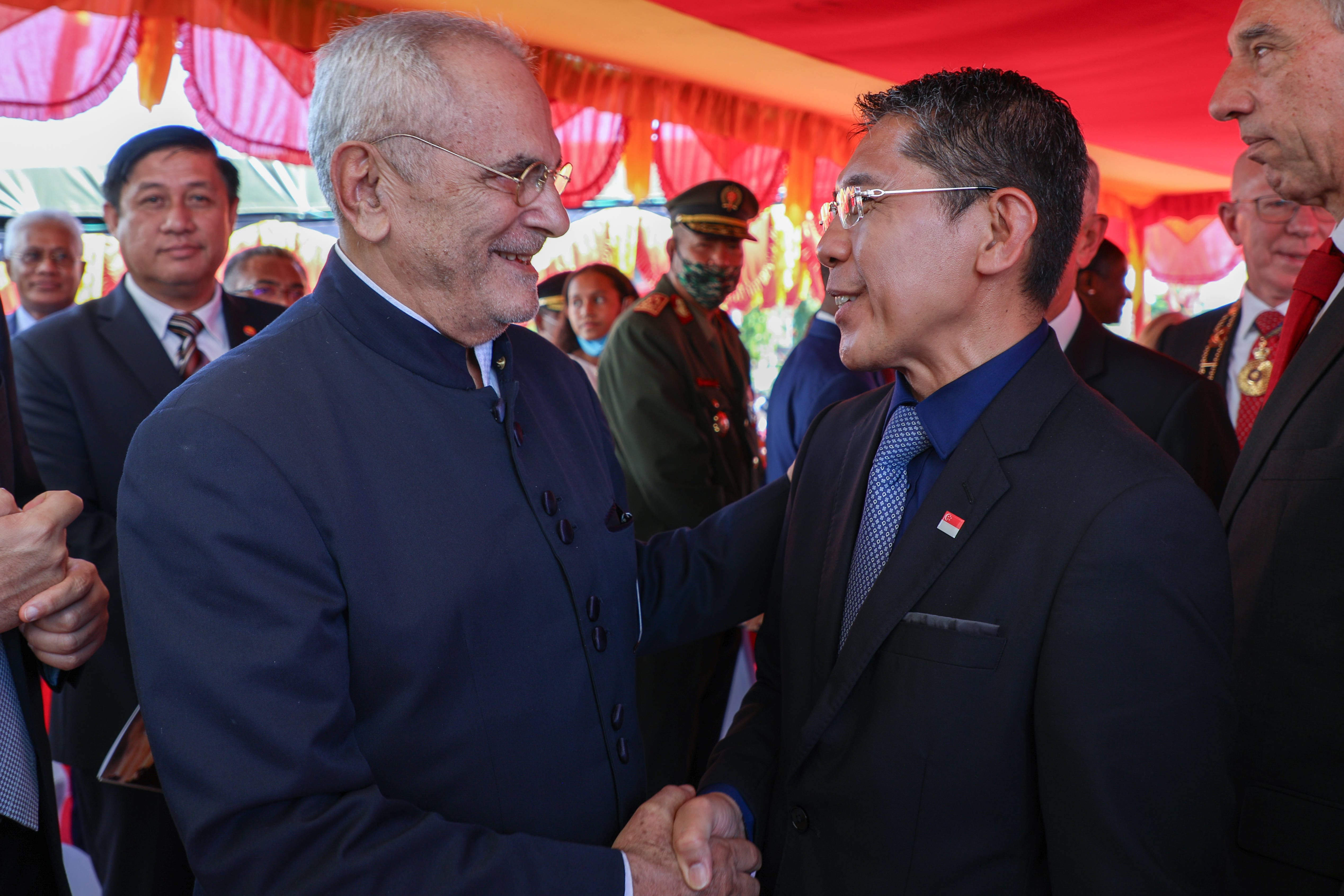 Two men in suits shake hands outdoors, one with a Singapore flag pin. Others stand nearby.