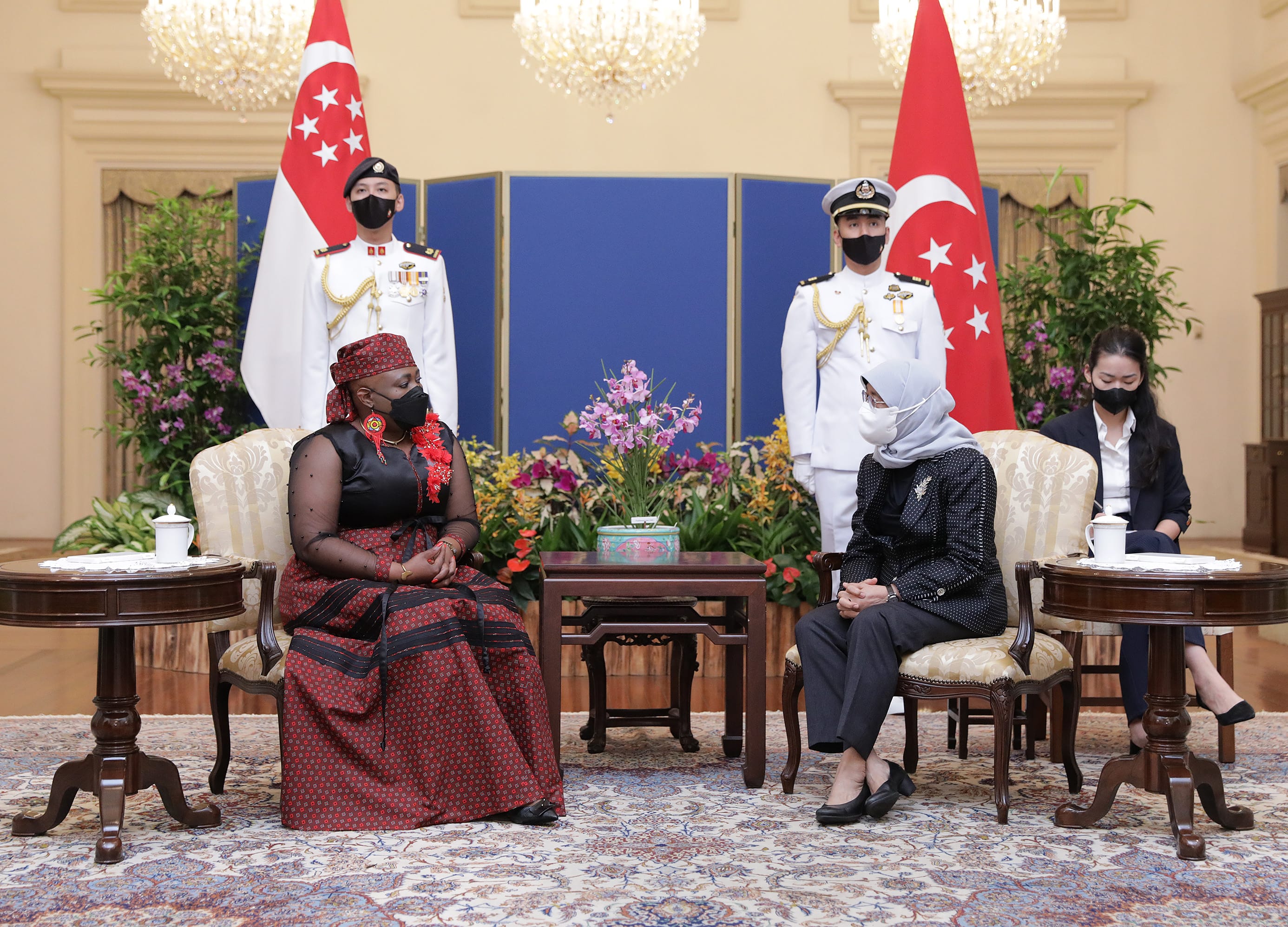Two women seated, with guards and Singapore flags behind, wearing face masks.