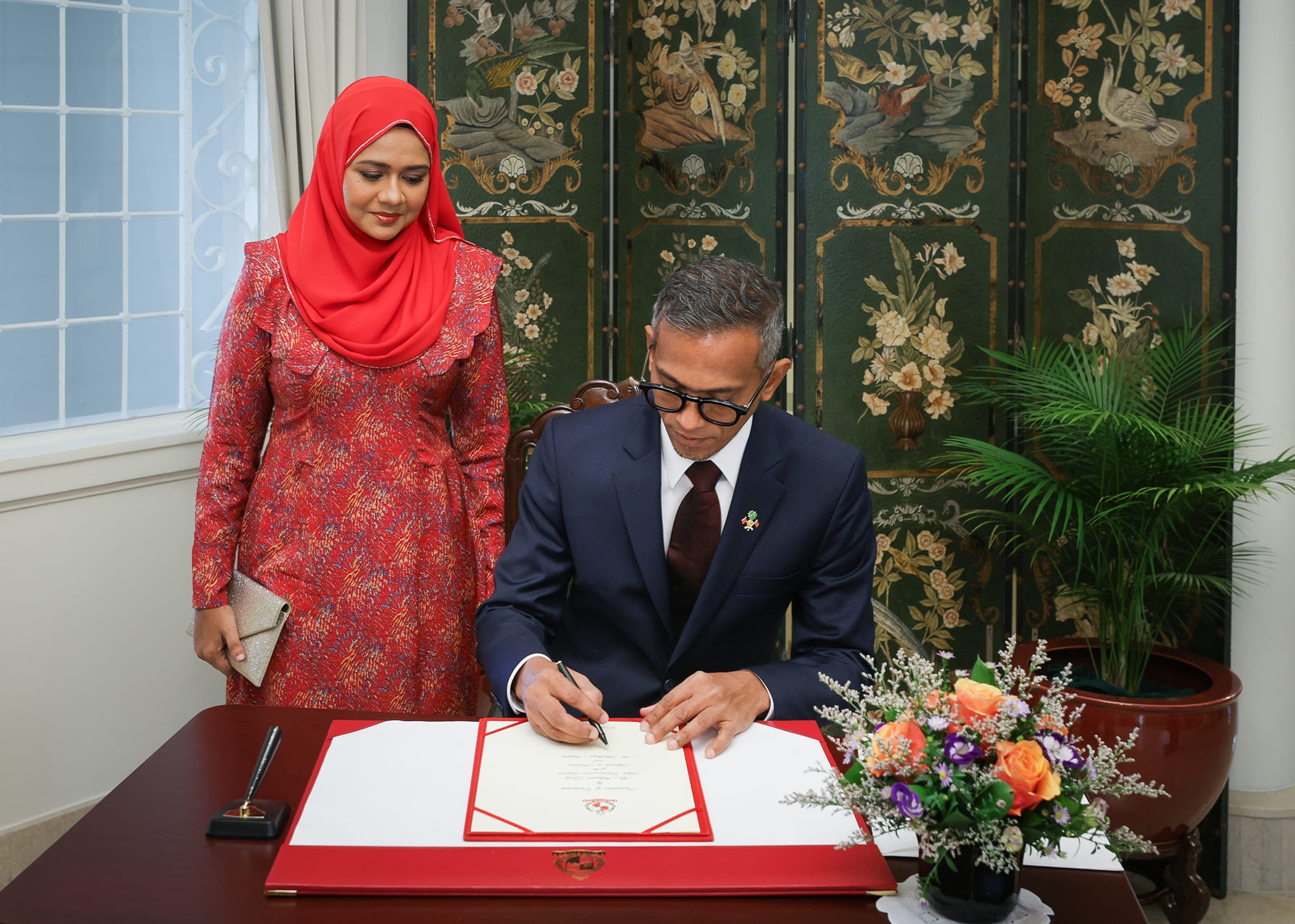 Man signing a document at a table as a woman in a red hijab stands beside him.