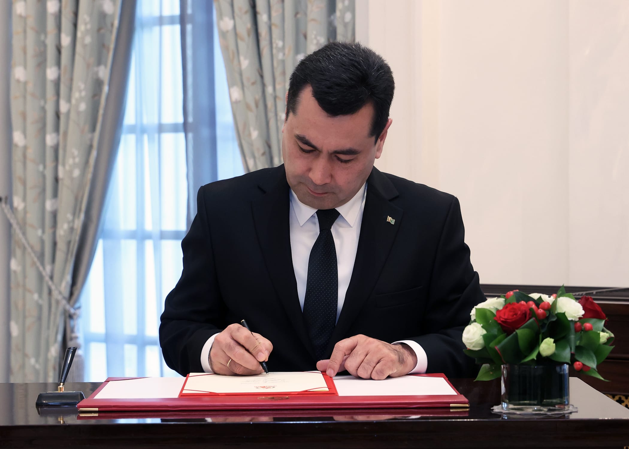 Man in suit signing document on red-trimmed desk with flowers and pen stand.