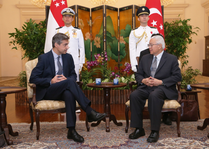 Two men in suits sitting, facing each other. Singaporean flags and guards stand behind them.