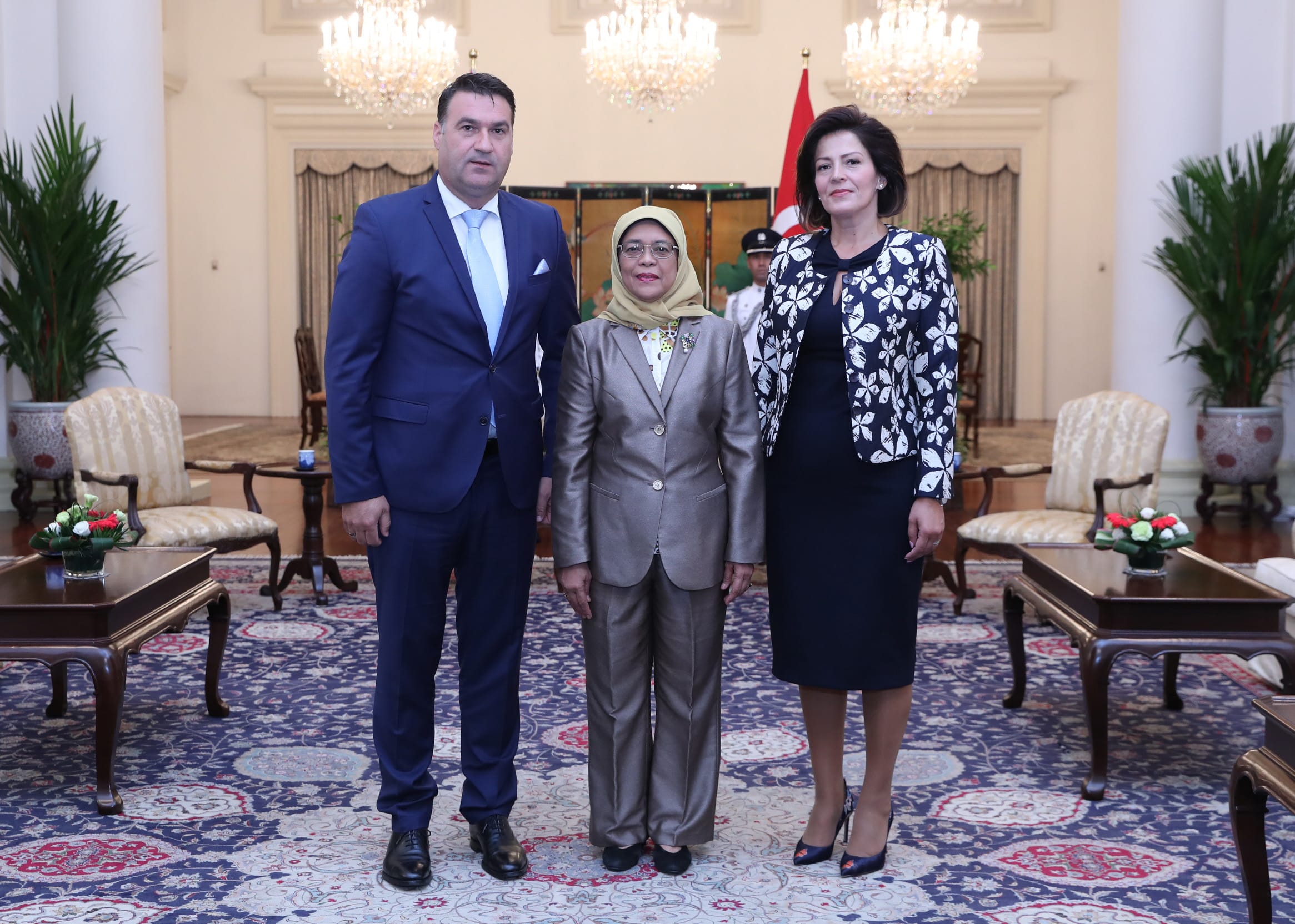 Three people stand in a room with chandeliers and a patterned rug; Singapore flag visible.