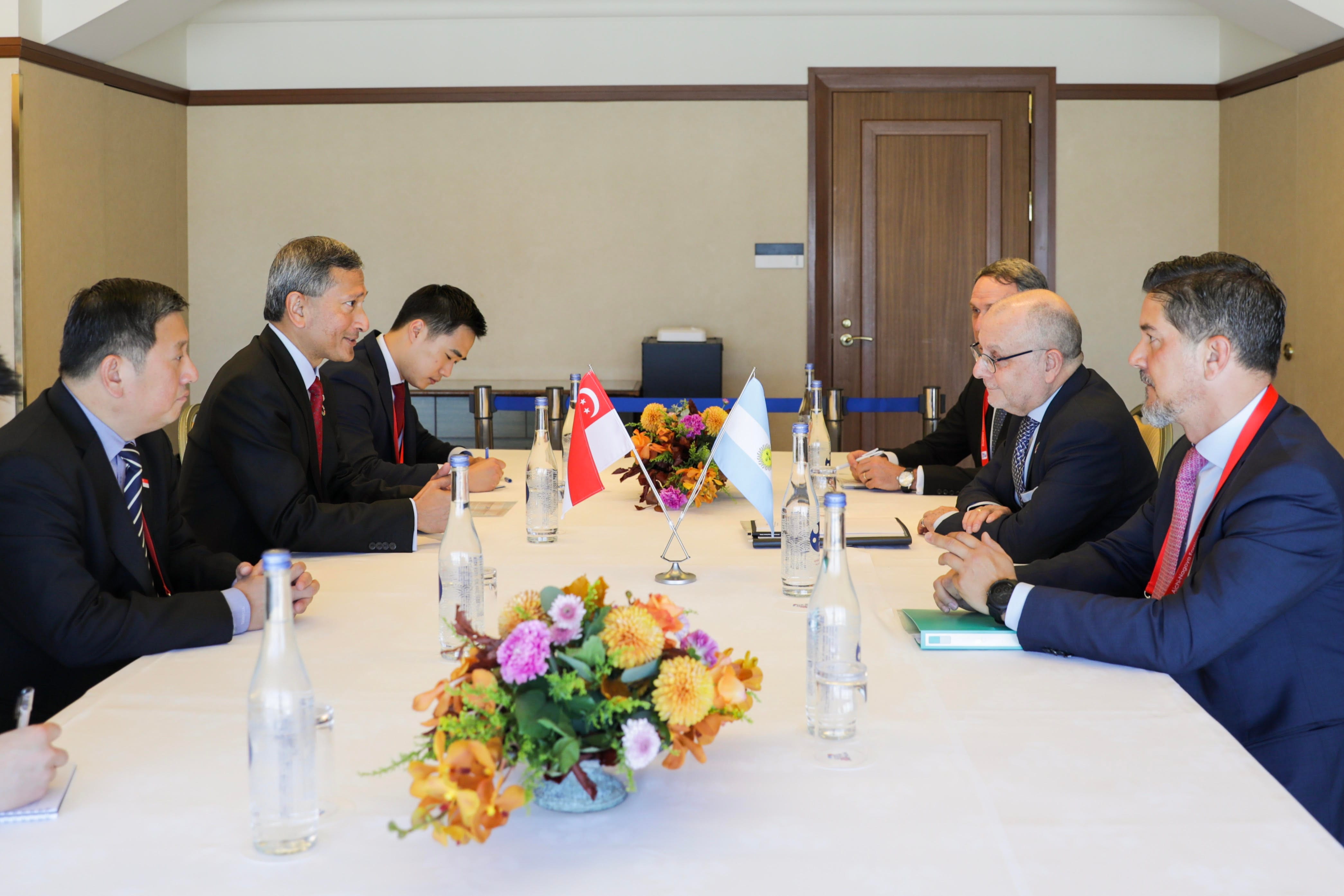 Meeting with men in suits at a long table with flags and floral arrangements.