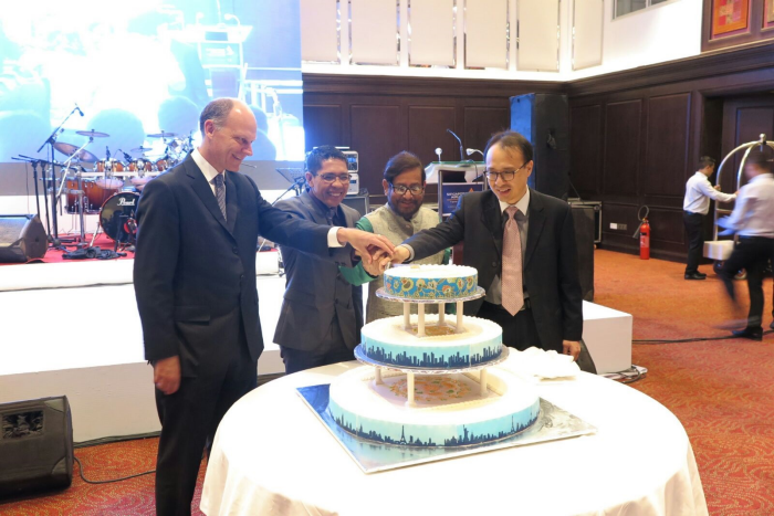 Four people in suits cut a tiered cake decorated with city skylines at an event.