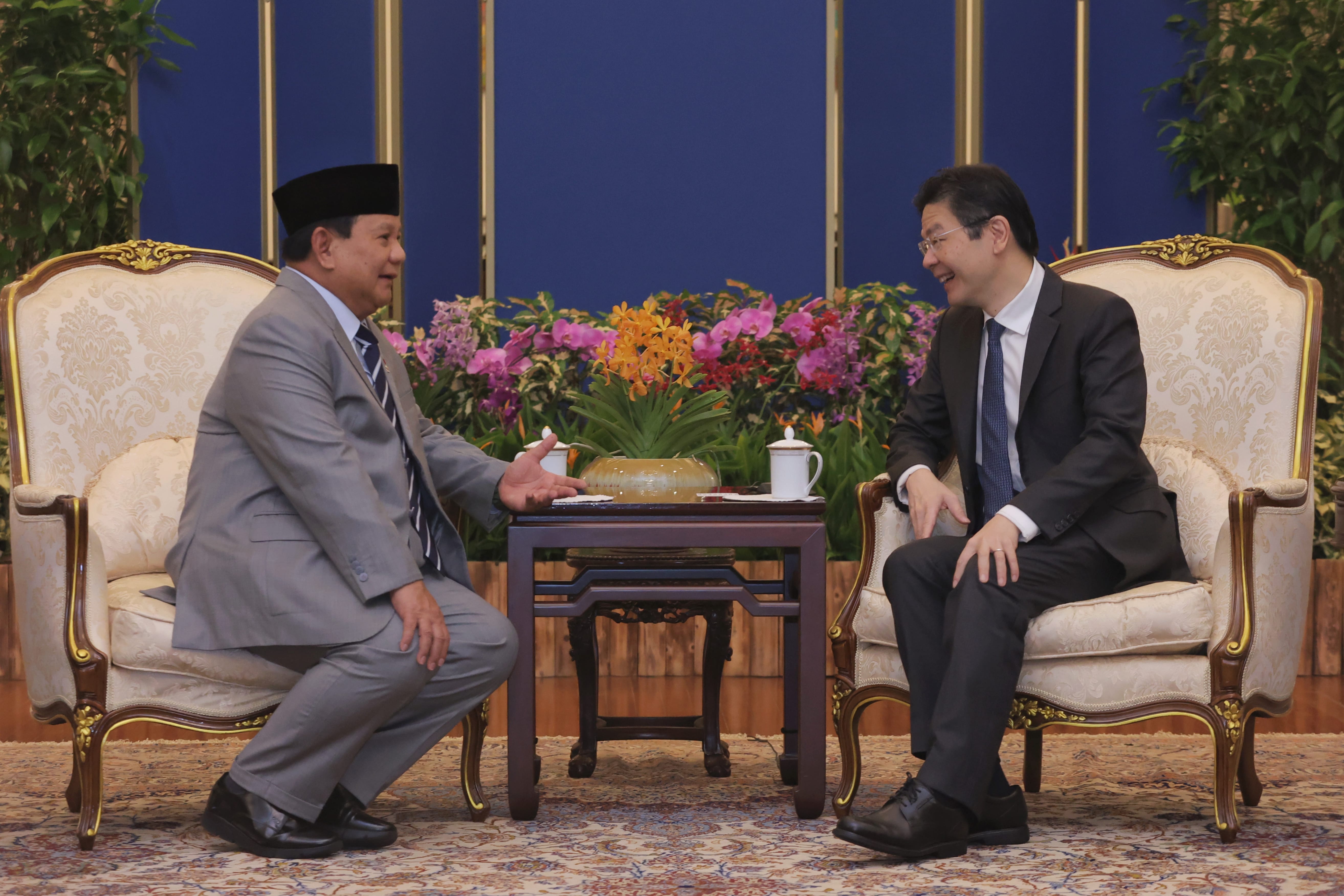 Two men in suits sit across from each other with a table between them.