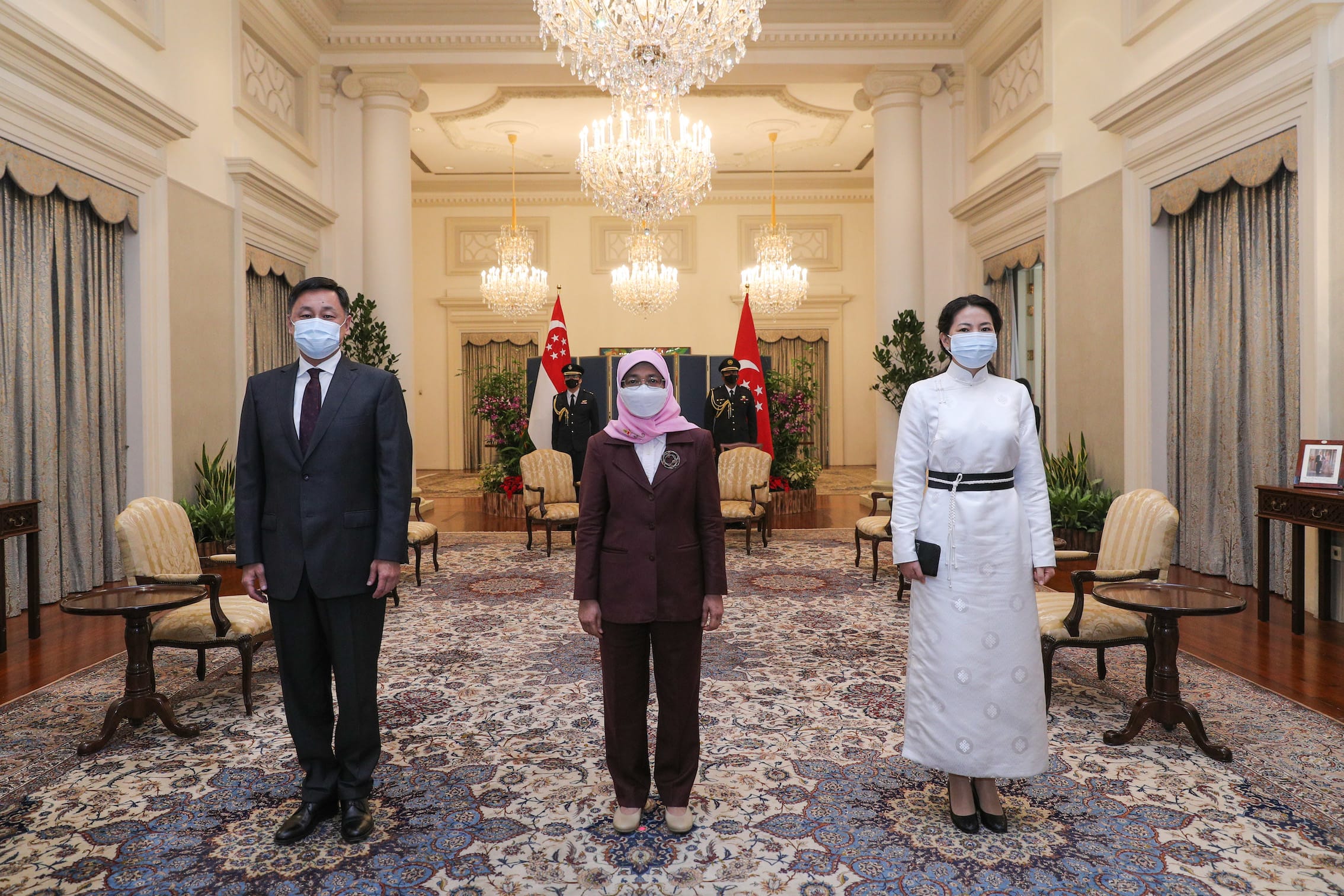 Three people in face masks stand inside a room with Singapore flags and chandeliers.